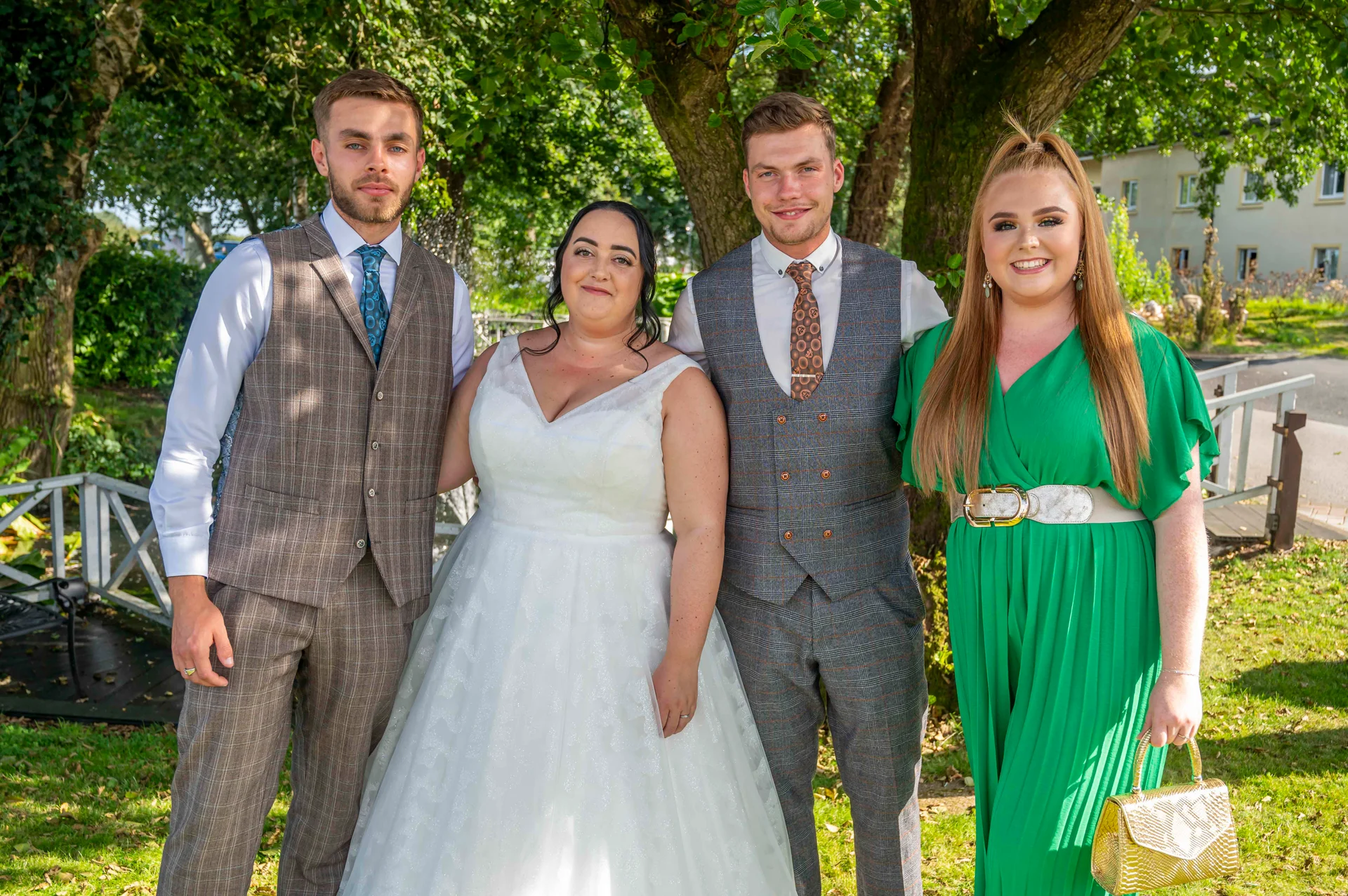 A bridal party of four poses outside on a sunny day next to mature trees and a white wooden fence.