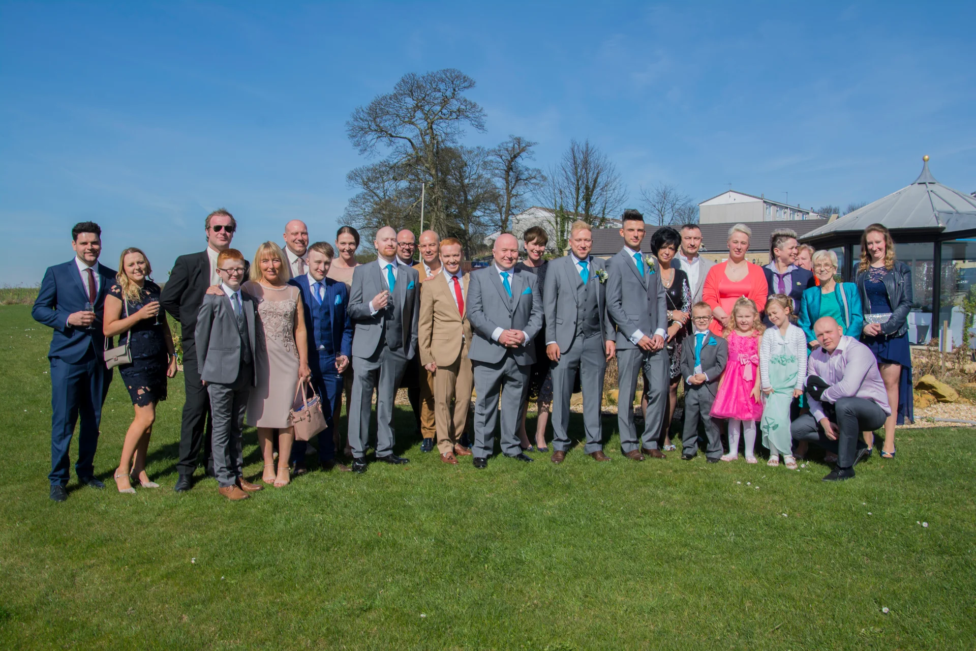 A large formal wedding group wearing suits and dresses poses together on a bright, sunlit lawn under a clear blue sky.