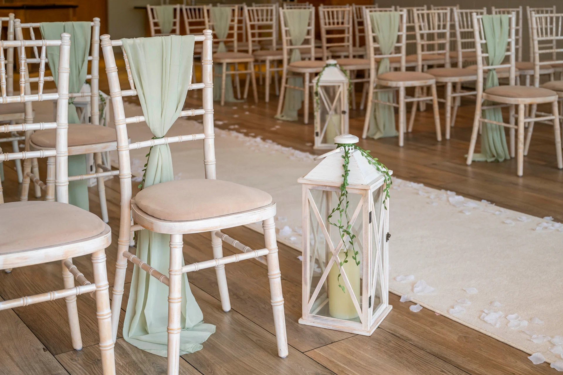 An empty wedding ceremony aisle is decorated with white lanterns, greenery, and scattered petals alongside rows of chairs draped in sage green fabric.
