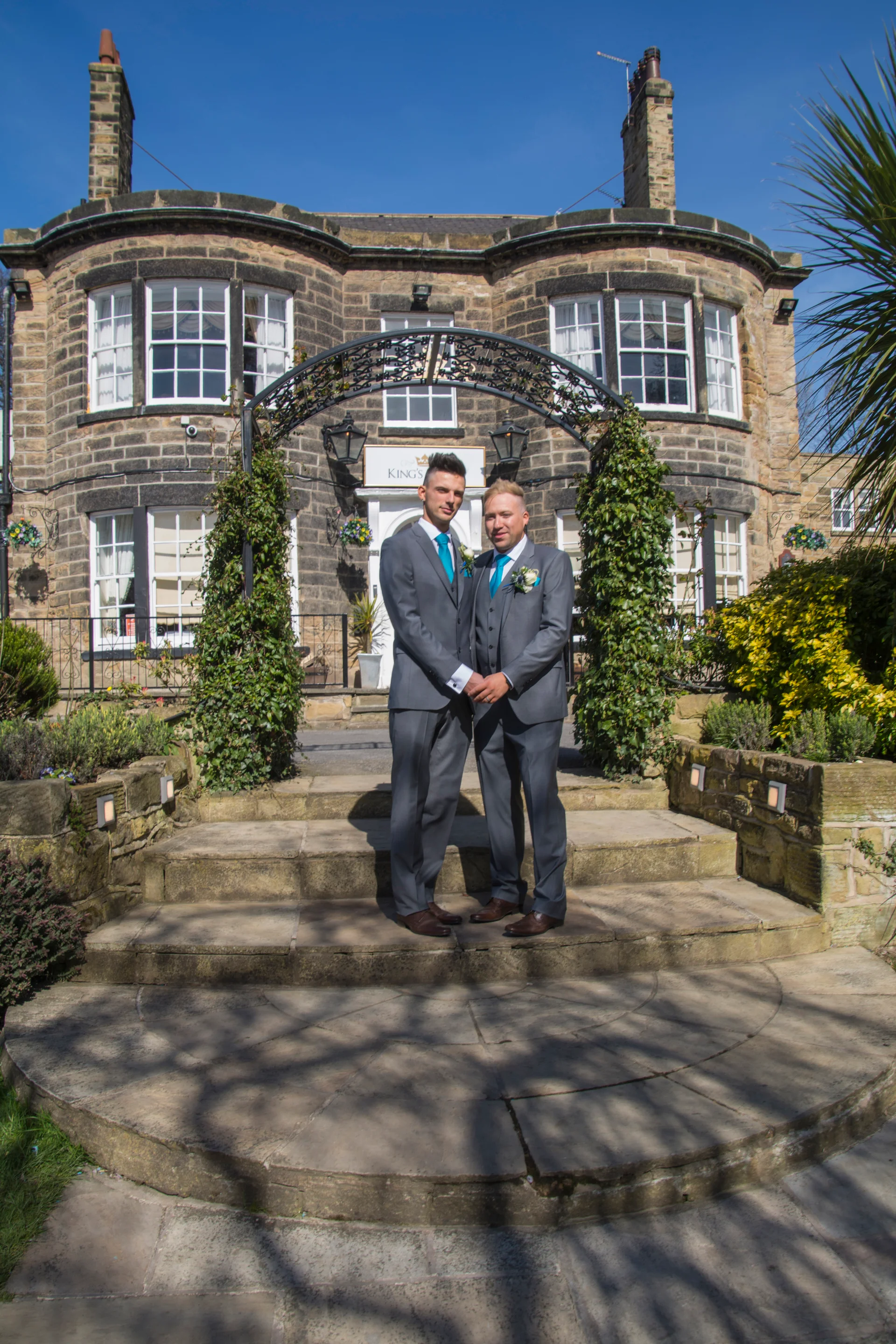 Two grooms in matching grey suits stand hand in hand under a vine-covered archway outside a stone hotel on a sunny day.