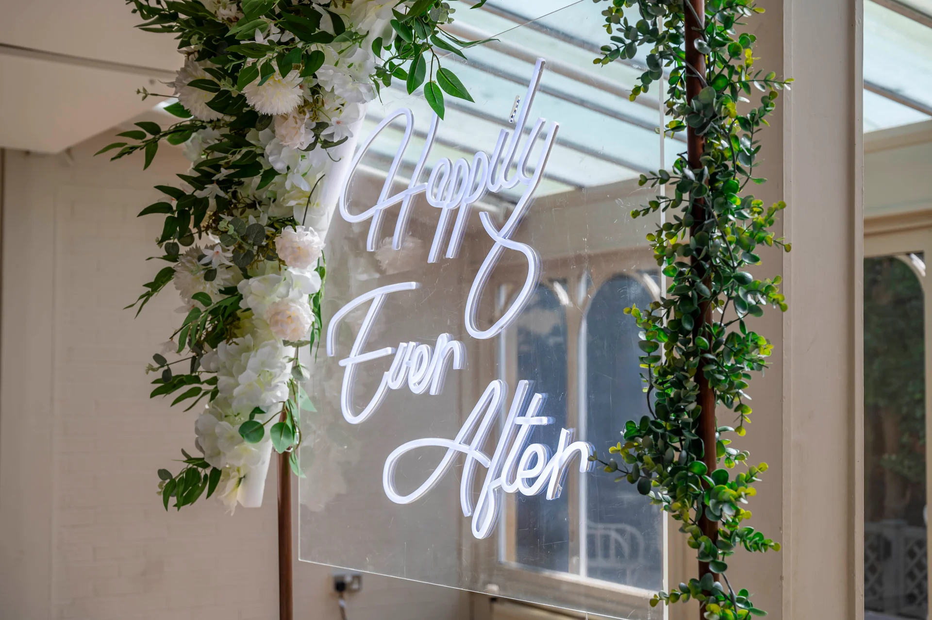 An illuminated acrylic neon sign displaying happily ever after is framed with lush white floral arrangements and green foliage inside a bright orangery venue.