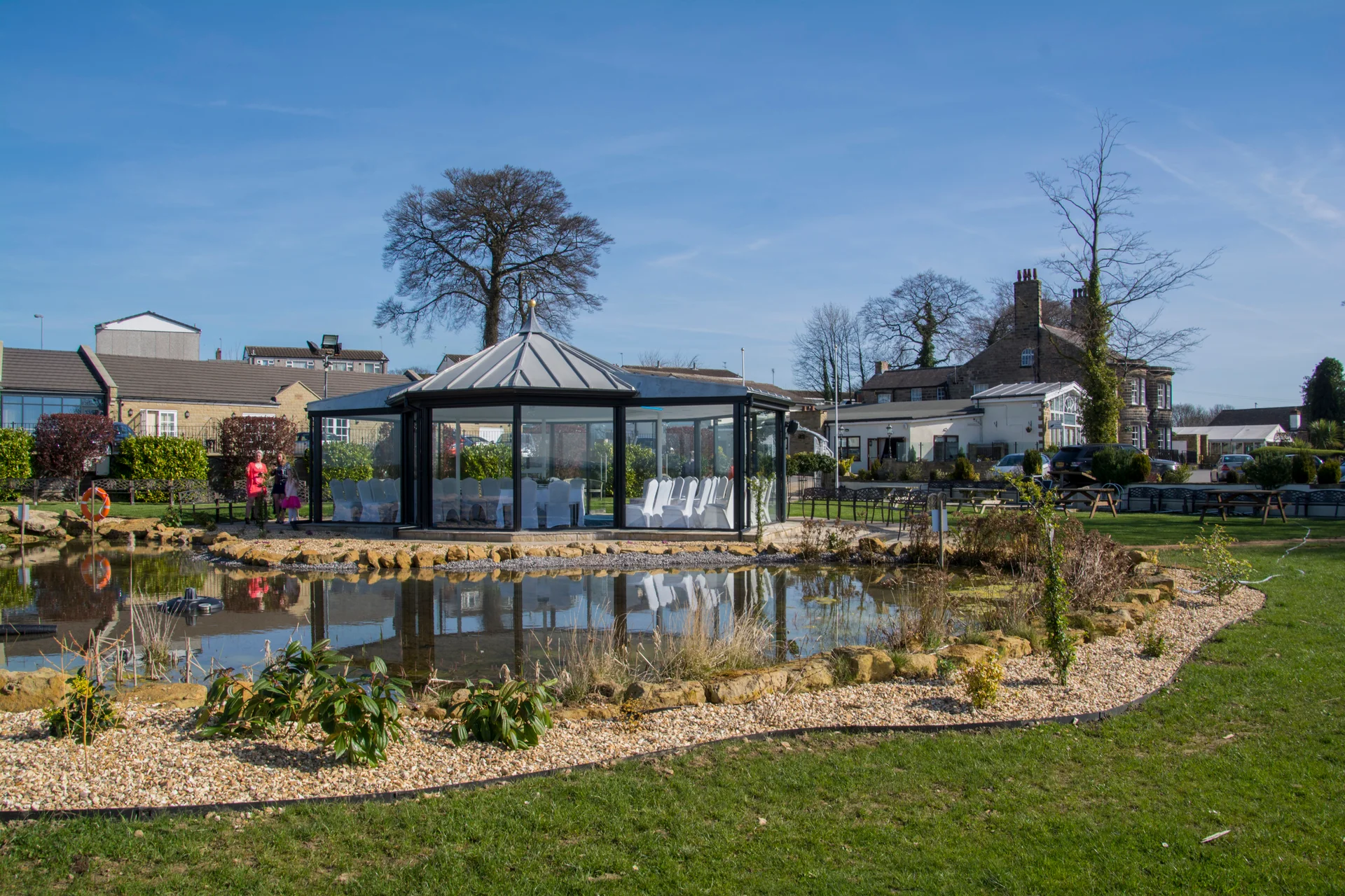 A modern glass wedding gazebo stands beside a tranquil pond under a clear blue sky on a sunny day.