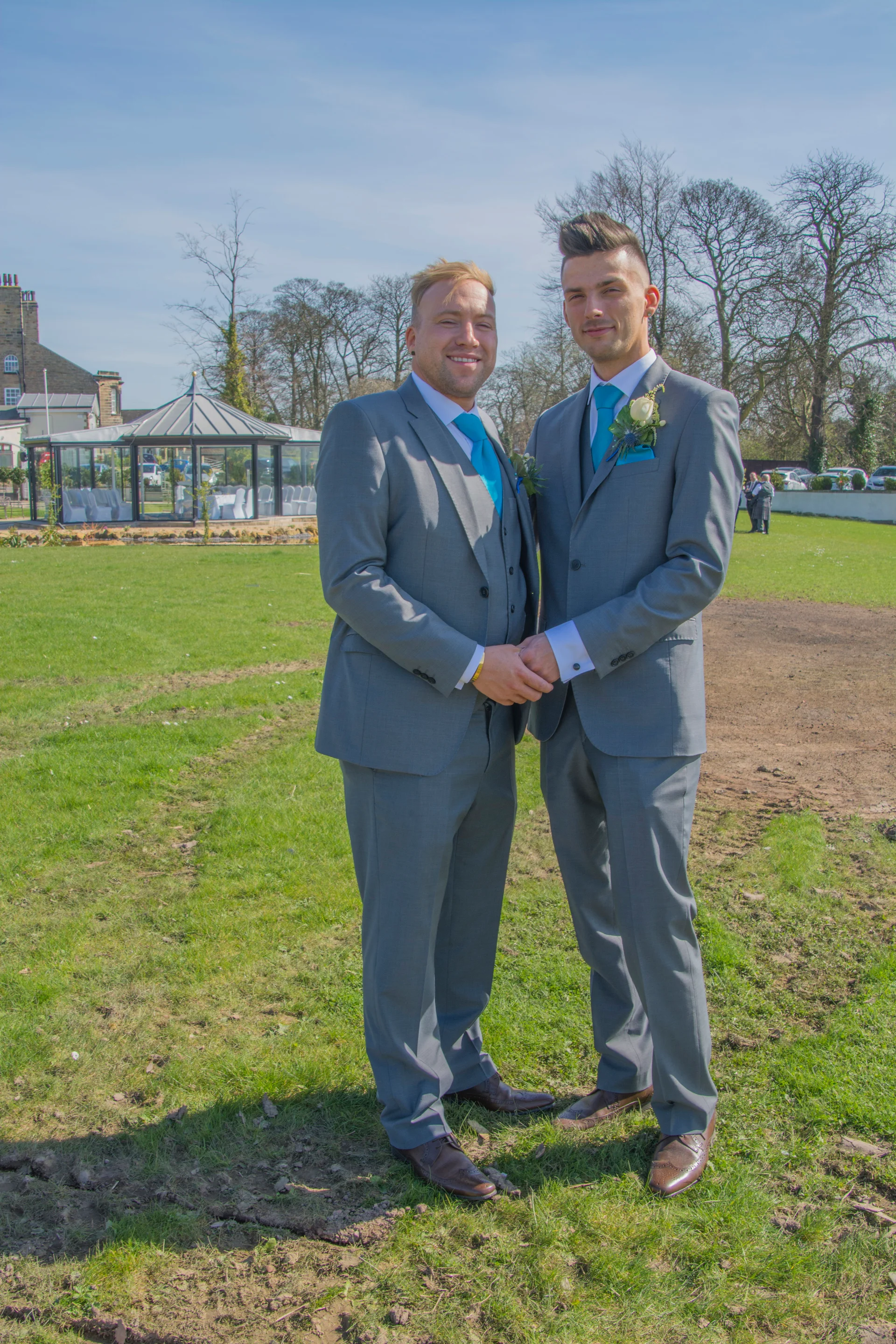 Two grooms in matching grey suits and bright blue ties stand hand in hand on a green lawn outside a wedding venue on a sunny day.