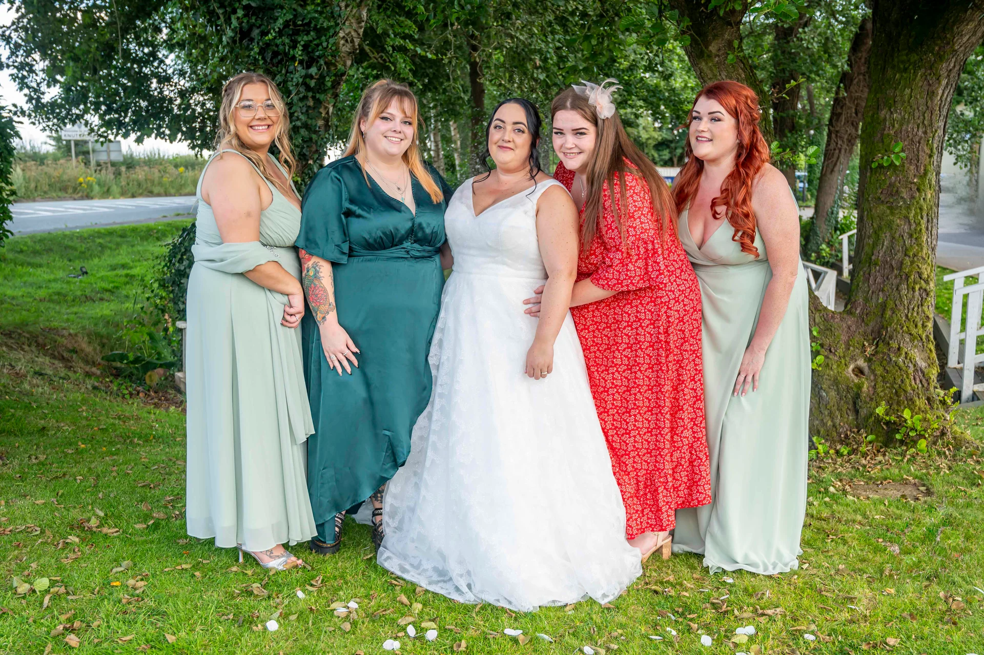 The bride in a white lace a-line gown stands outdoors with her four bridesmaids wearing mismatched sage green and red dresses against a lush garden backdrop.