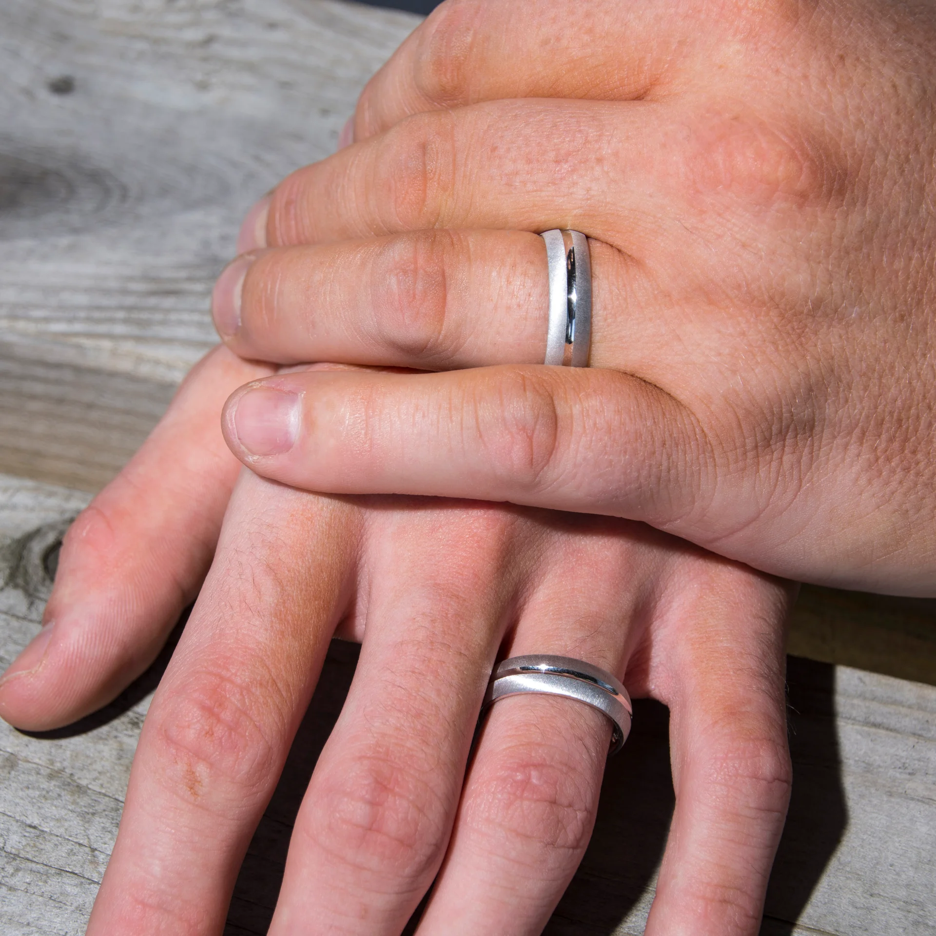 Close-up of two grooms holding hands, showcasing their matching silver wedding bands against a textured wooden background.