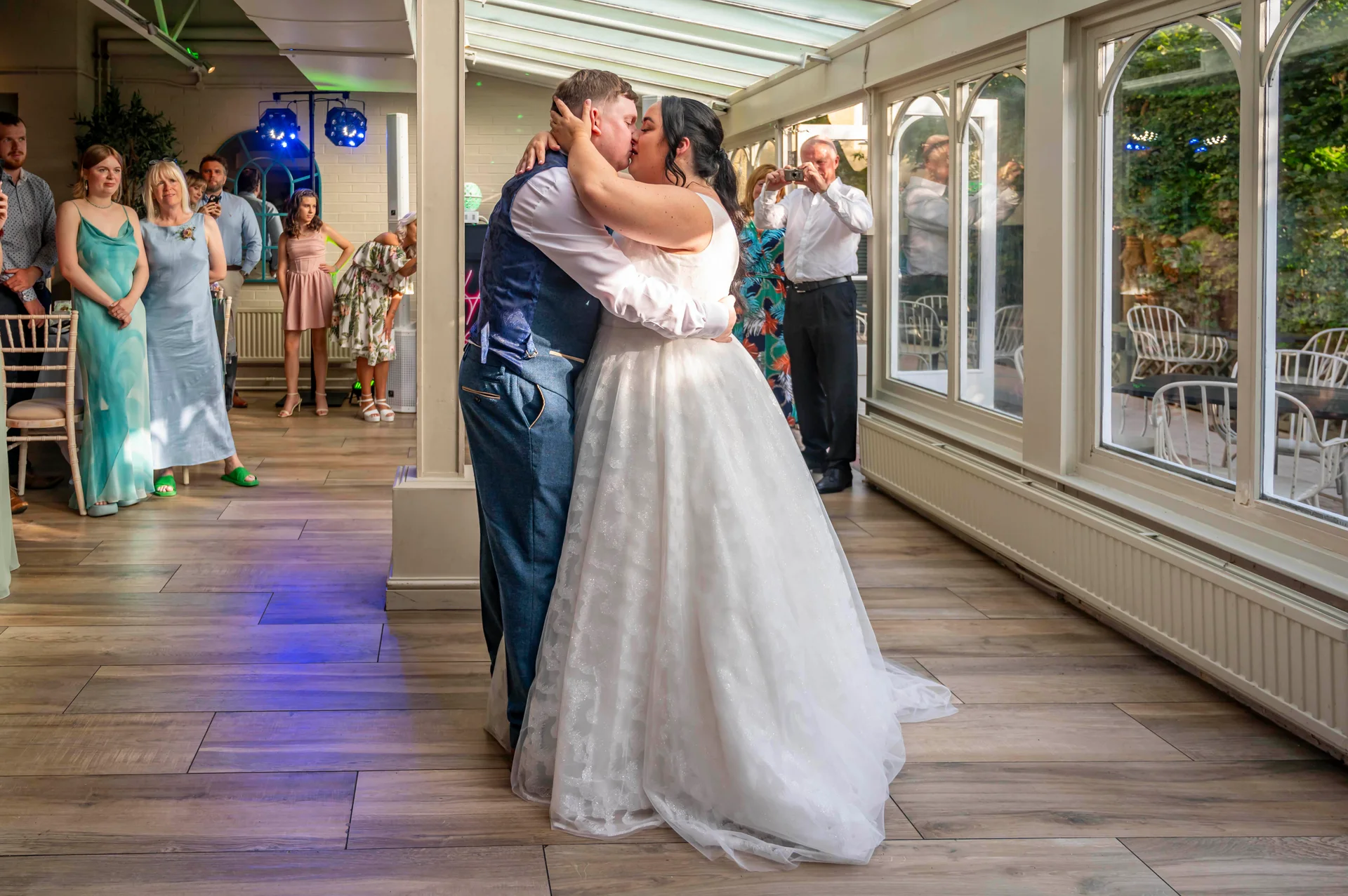 The newly married couple share an intimate first kiss on the dance floor inside a light-filled conservatory while wedding guests watch and photograph them.
