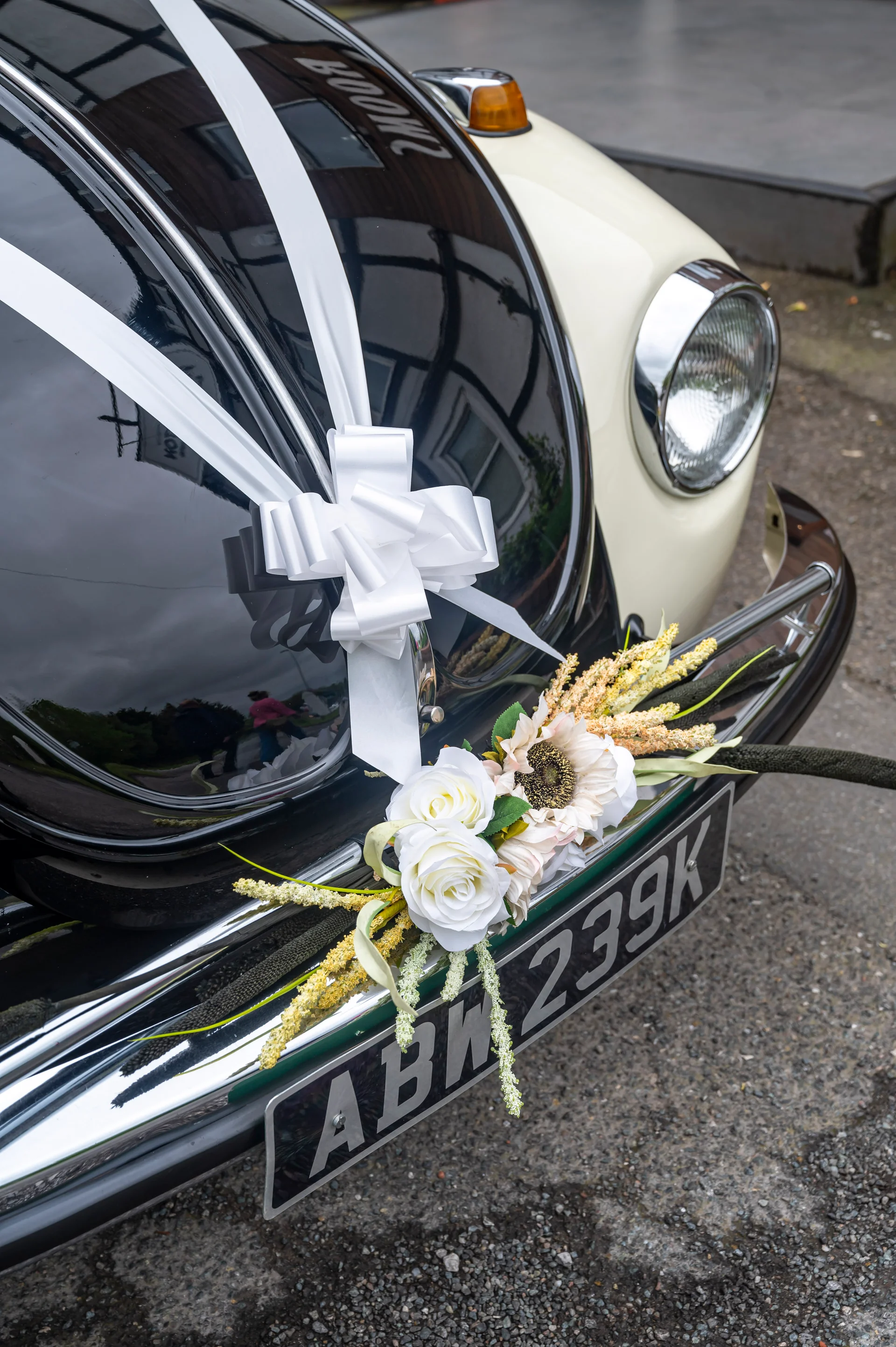 A vintage Volkswagen Beetle wedding car decorated with a white ribbon bow and floral arrangement on the front bumper.