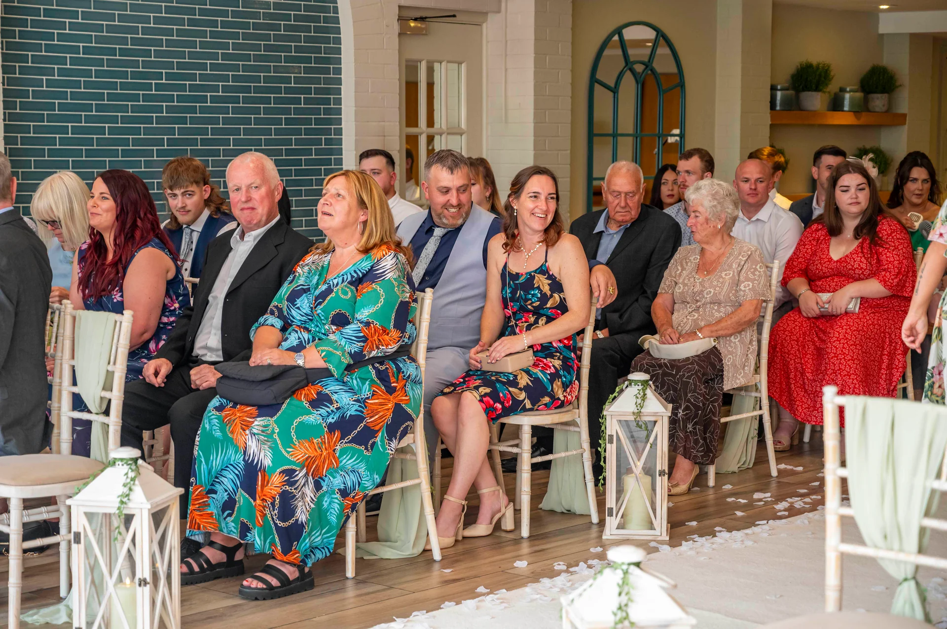 A candid wide shot captures wedding guests seated on chairs lining the aisle inside a brightly lit venue adorned with white lanterns and scattered petals.