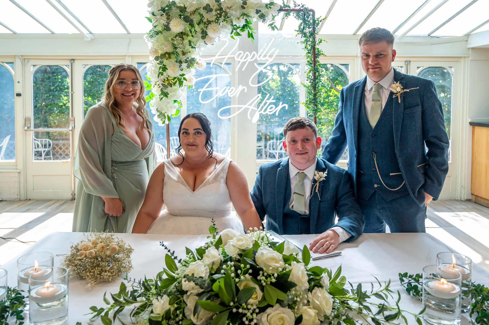 The newly married couple sits together at a white-clothed table to sign their register, flanked by a bridesmaid and groomsman in an airy orangery venue decorated with a glowing neon sign and abundant white and green florals.