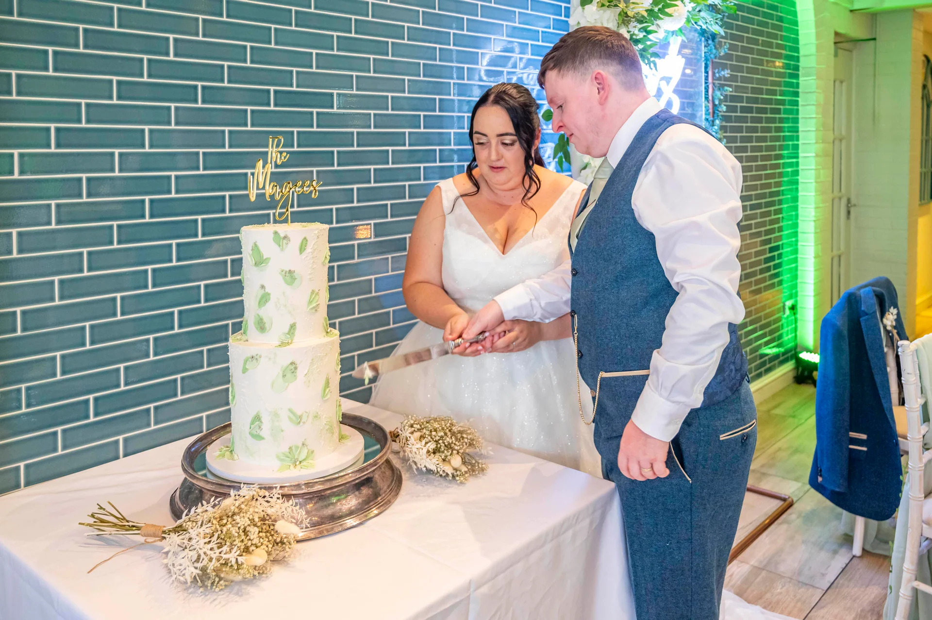A newlywed couple smiling as they cut a two-tier white buttercream wedding cake decorated with painted green leaf patterns against a teal tiled wall.