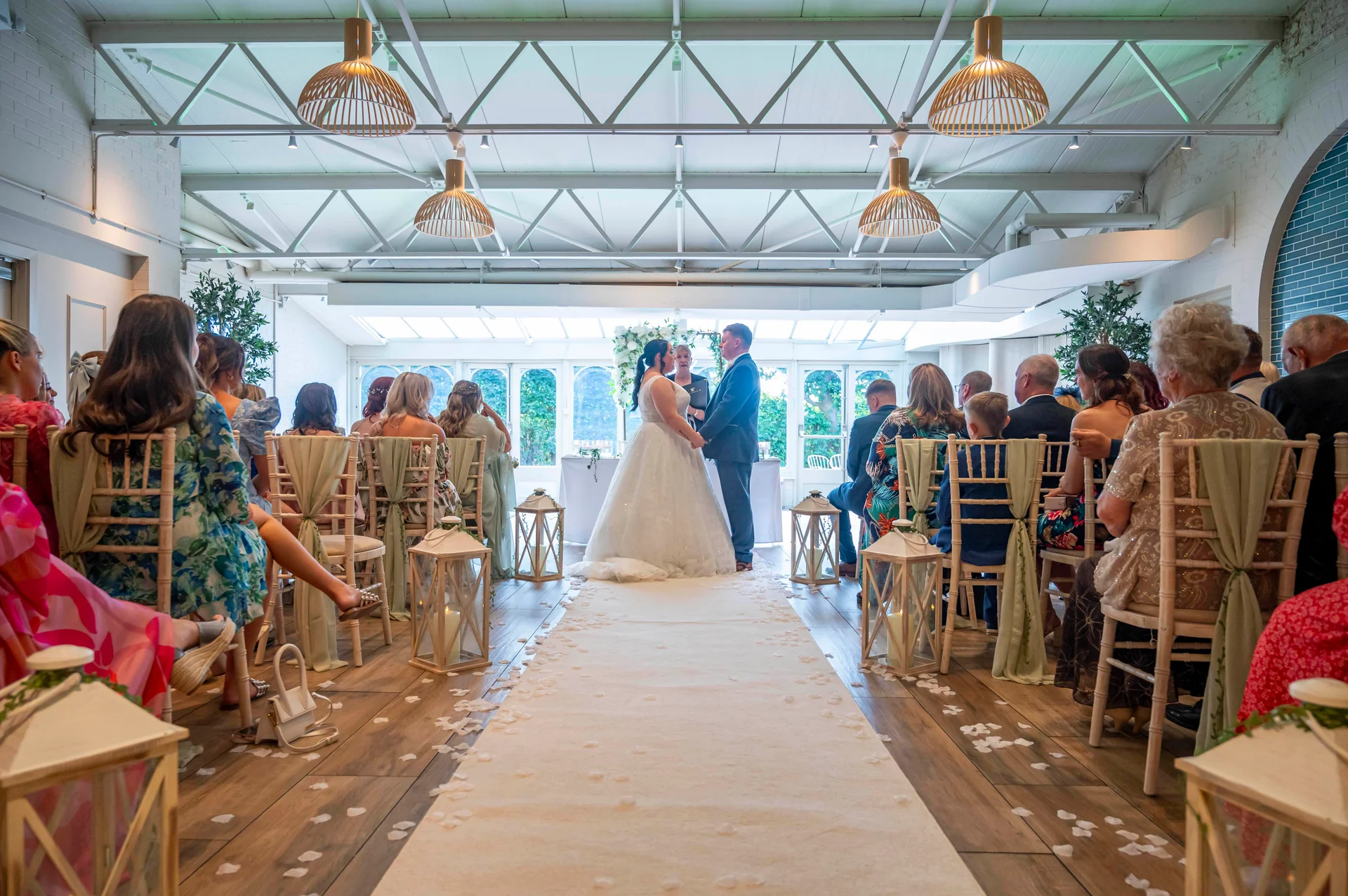A wide angle interior shot of an industrial venue during a civil ceremony, featuring the bride and groom standing at the end of a petal-strewn aisle lined with lanterns and seated guests.