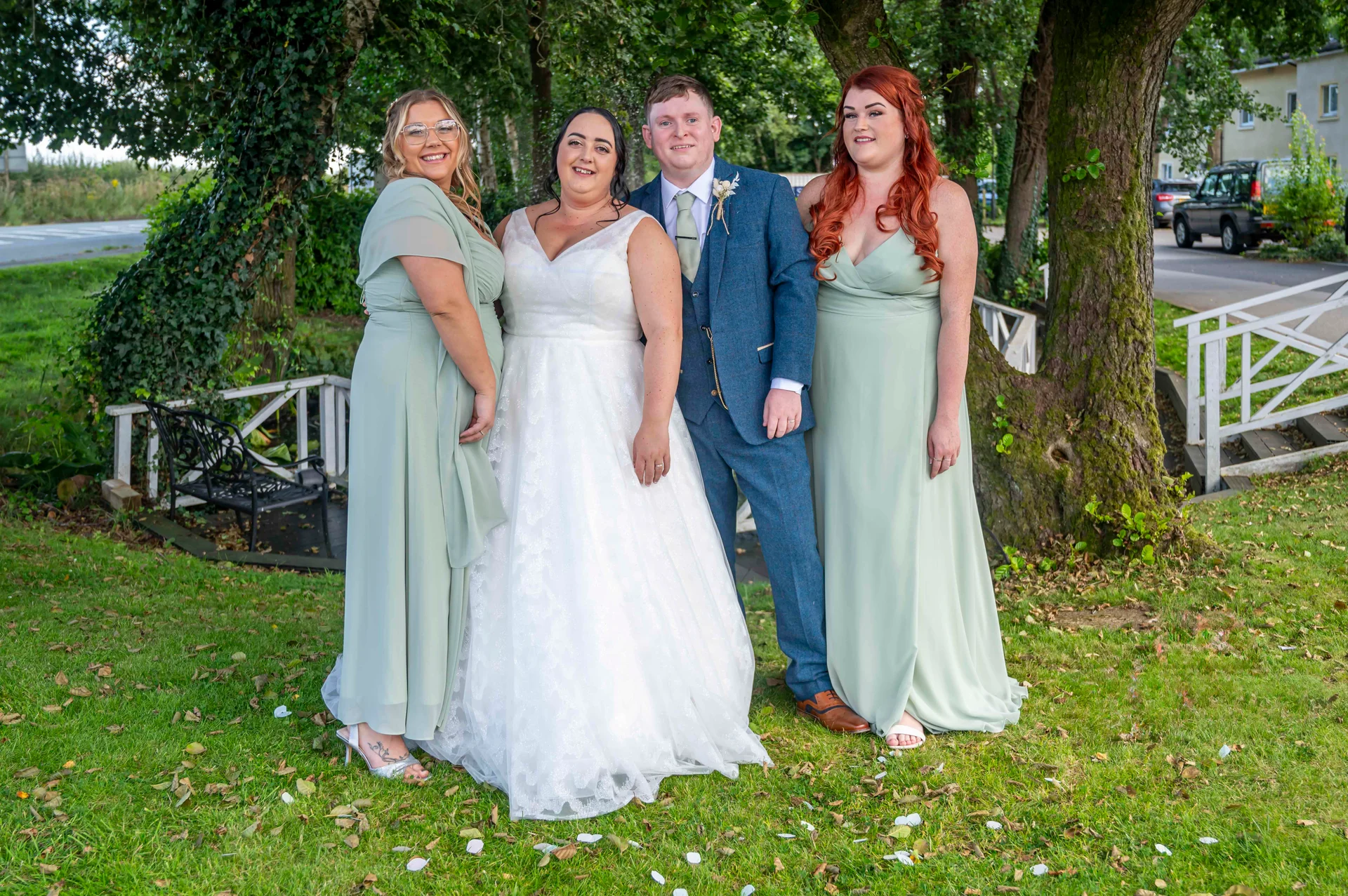 A newlywed couple poses outdoors on a grassy lawn with two bridesmaids wearing sage green dresses beside mature trees.