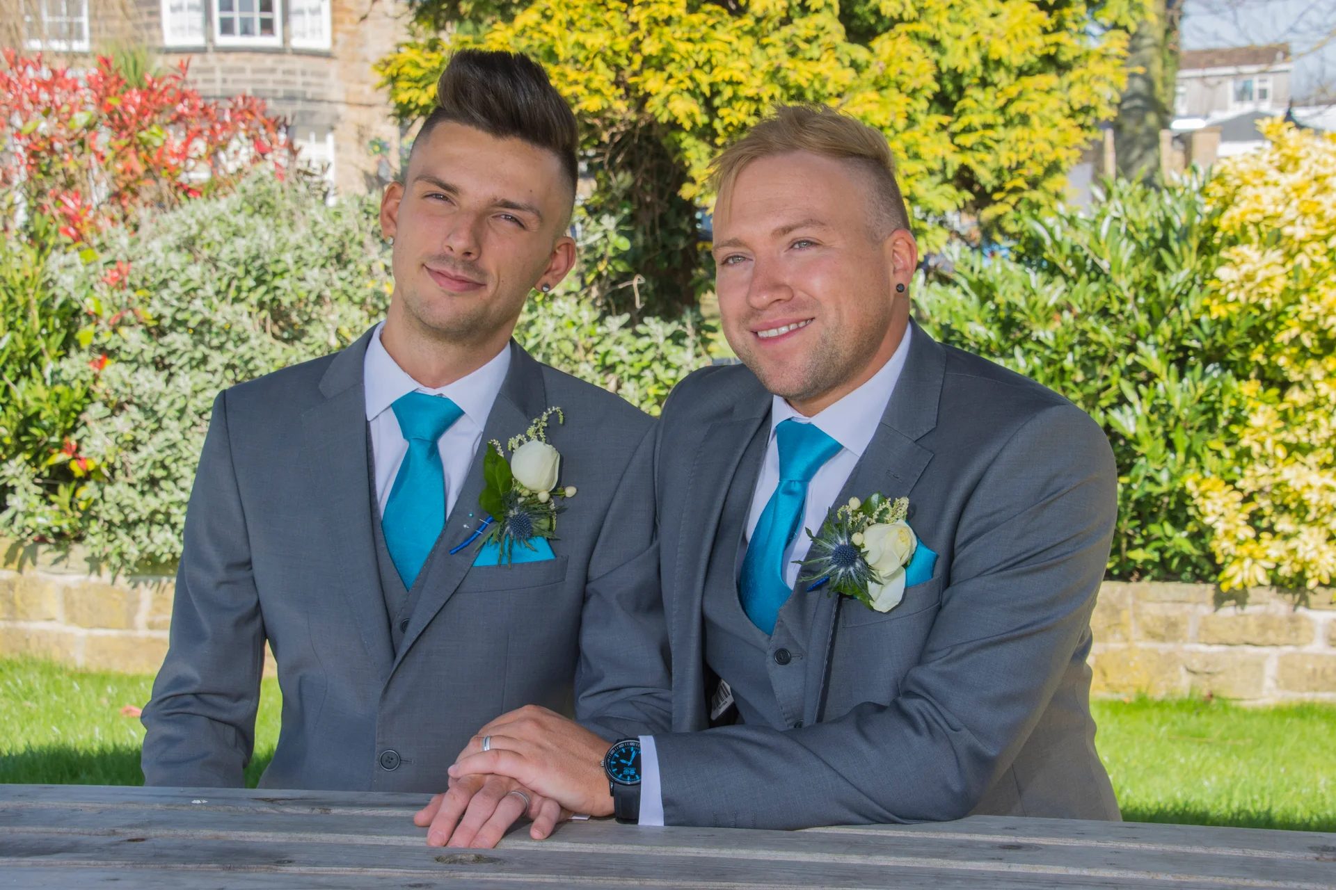 Two grooms in matching grey suits and bright blue ties pose together outdoors in a sunny garden, showcasing their wedding rings and boutonniere details.