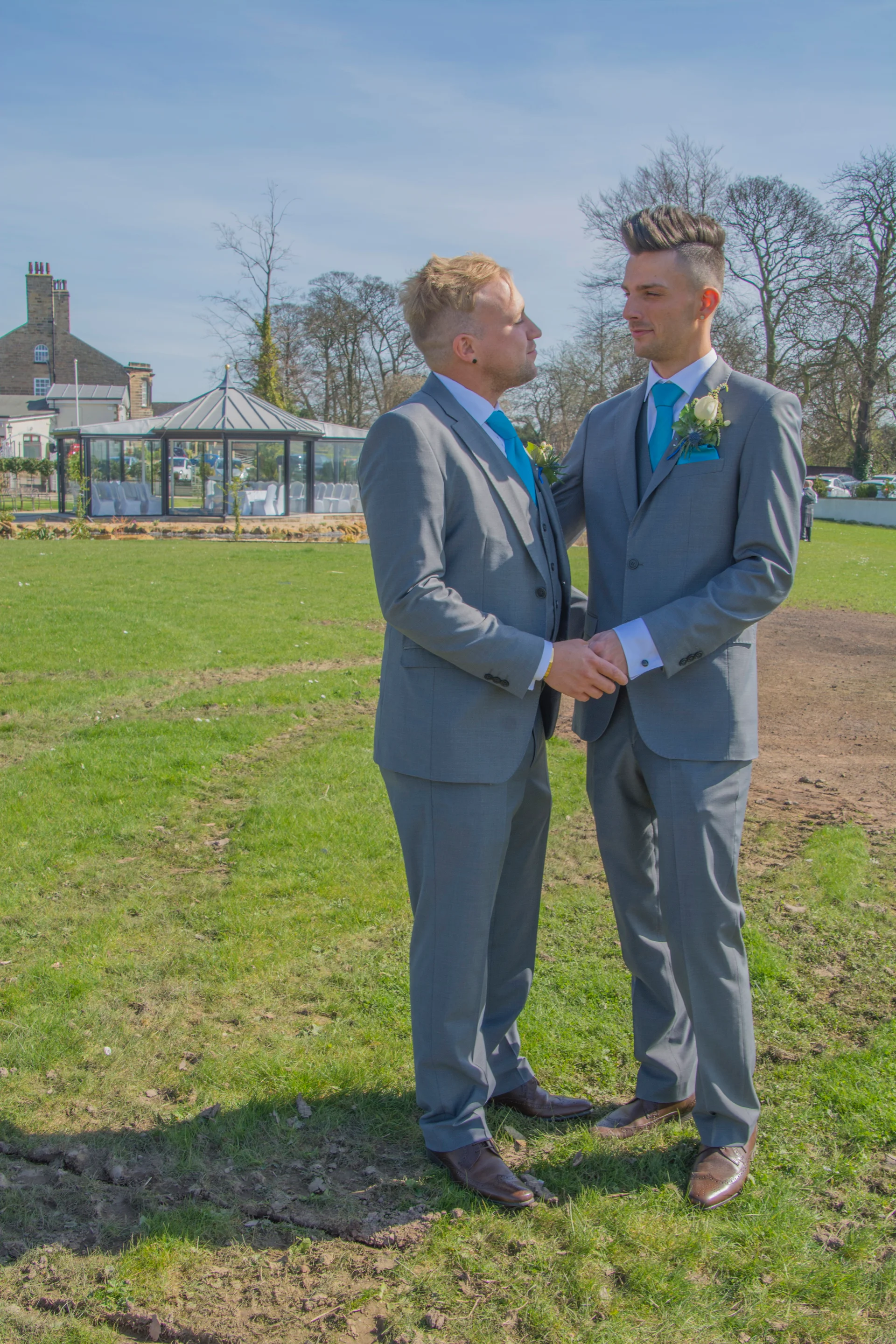 Two grooms in matching grey suits and turquoise ties hold hands outdoors on a grassy lawn with a glass conservatory in the background.