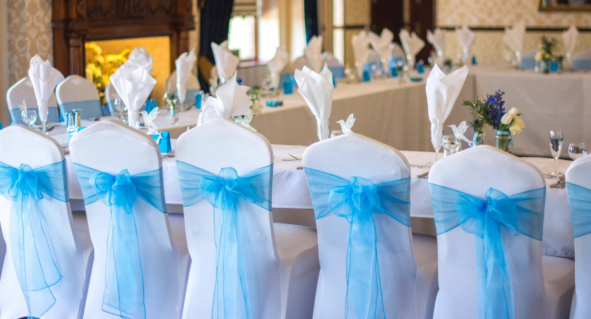 An elegant wedding breakfast reception setup featuring long banquet tables adorned with white linens and chairs decorated with bright blue organza sashes.