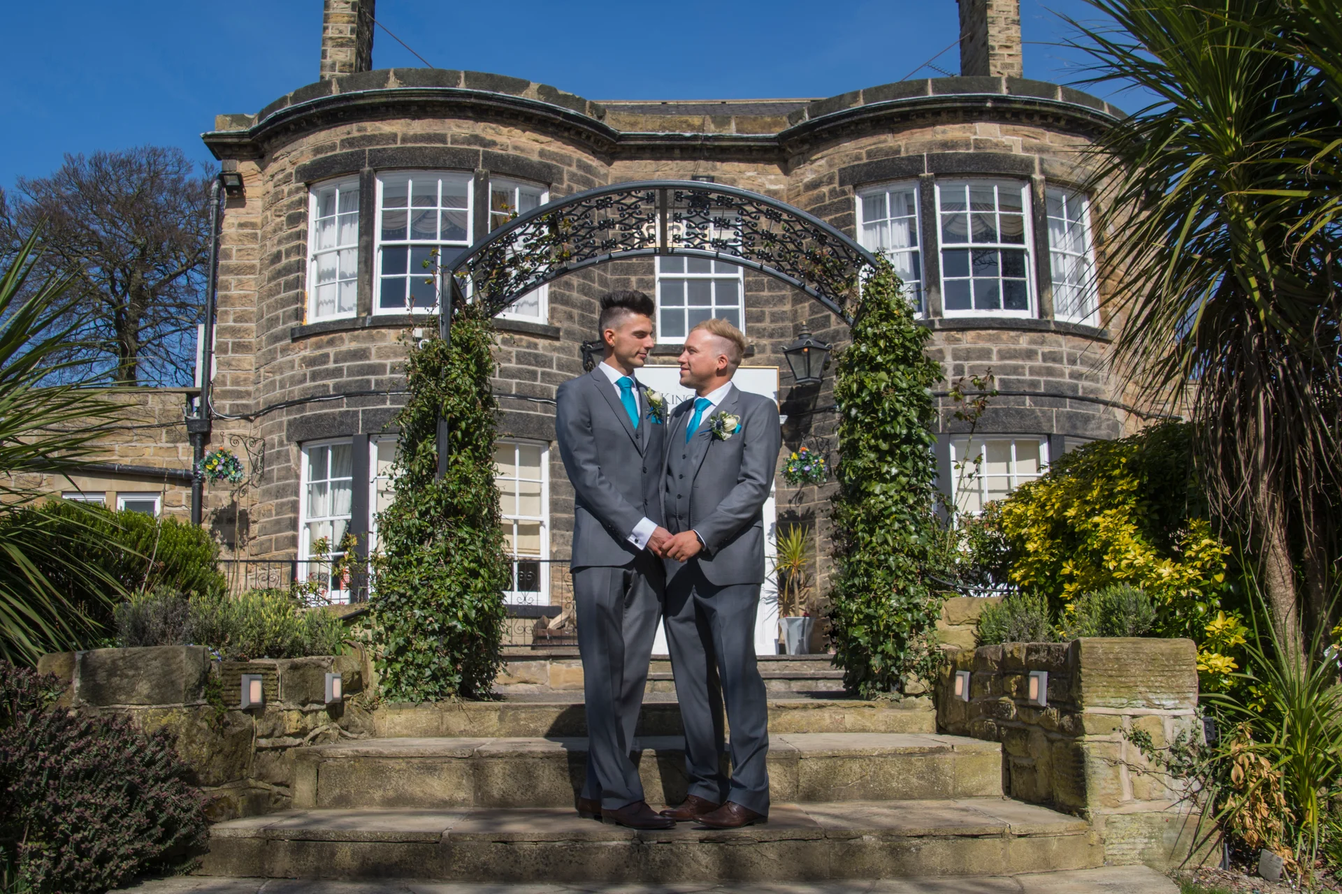Two grooms stand hand in hand beneath a floral archway outside a grand stone country house on a bright sunny day.