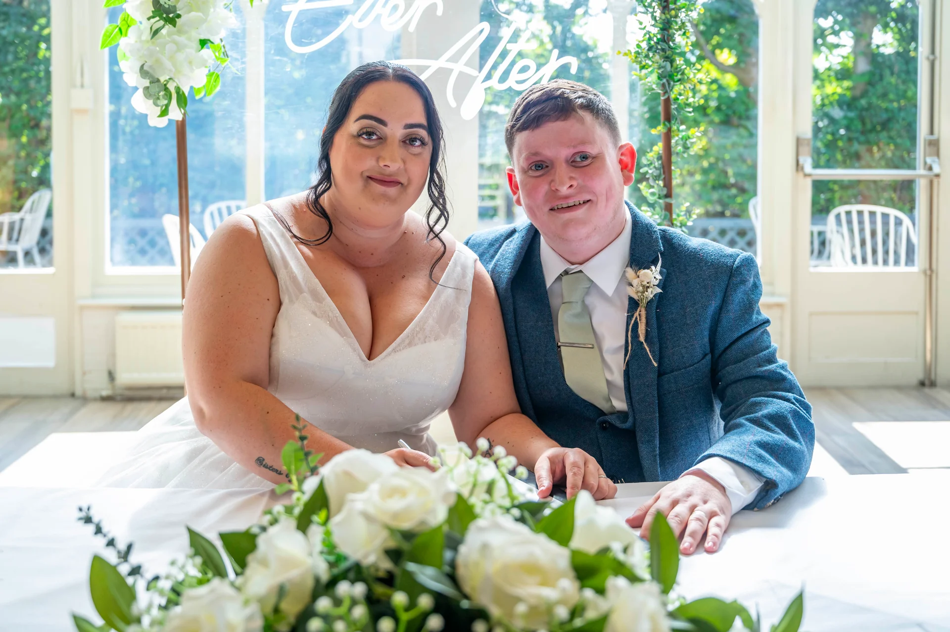 A smiling bride and groom sit together at a table in a light-filled venue, signing the register behind a display of white roses.