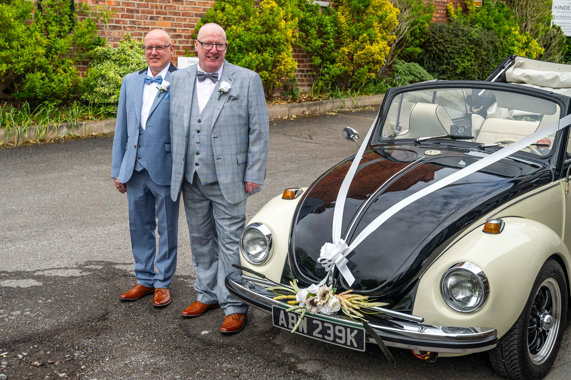 Two smiling grooms in matching grey checked three-piece suits pose proudly next to a vintage black and cream VW Beetle decorated with white ribbons and floral arrangements.
