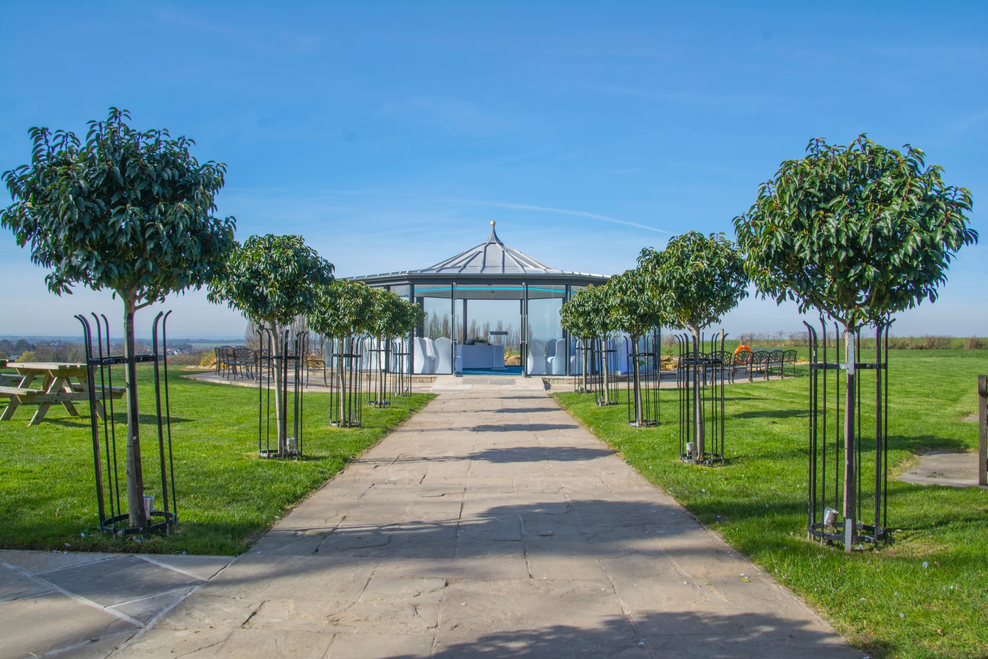 A stone paved path leads through manicured lawns towards a glass gazebo ceremony structure under a bright blue sky on a sunny day.