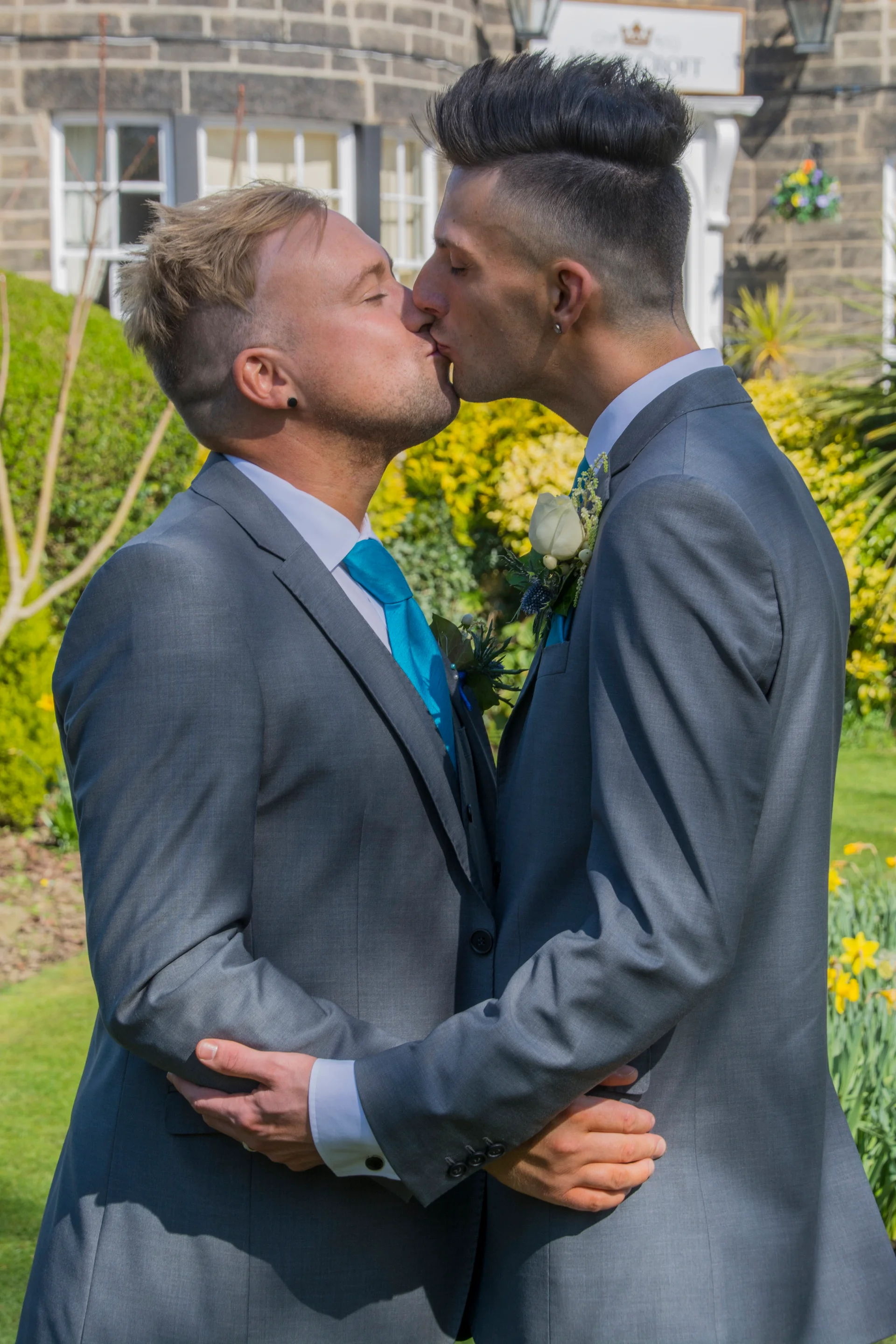 Two grooms share a tender first kiss outdoors in front of a stone building, both wearing matching grey suits and bright blue ties with white buttonholes.