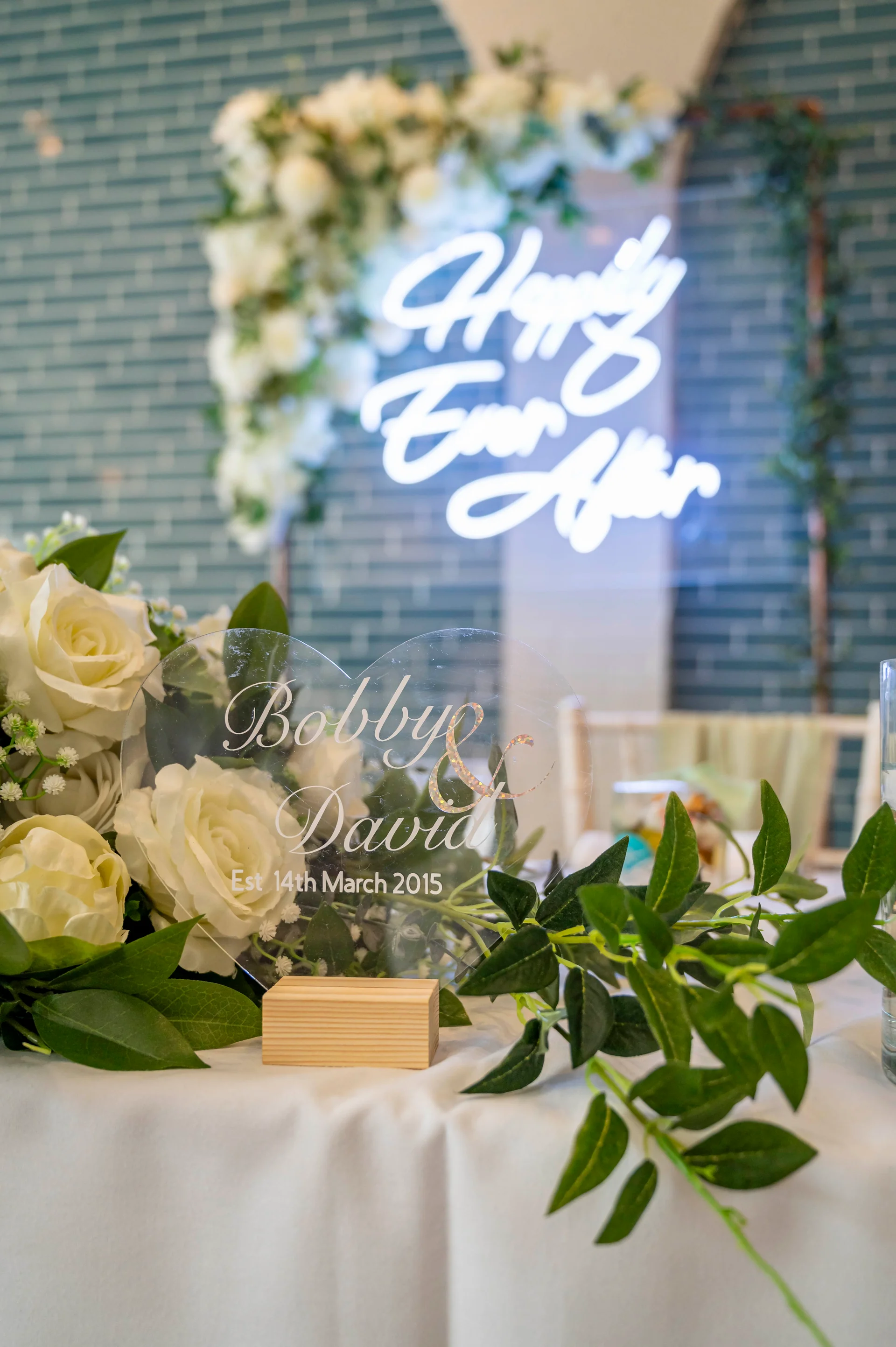 A close up view of a wedding reception tablescape featuring a heart-shaped acrylic sign with names, surrounded by white roses and greenery, with a glowing neon sign and floral arch backdrop against an exposed brick wall.