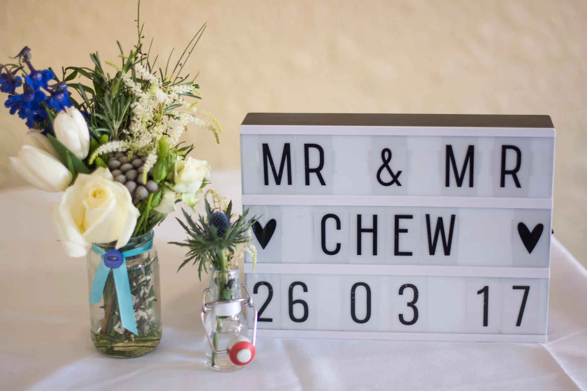 A light box displaying "Mr & Mr Chew 26 03 17" sits on a white table next to a jar of white roses, blue delphiniums, and thistles.