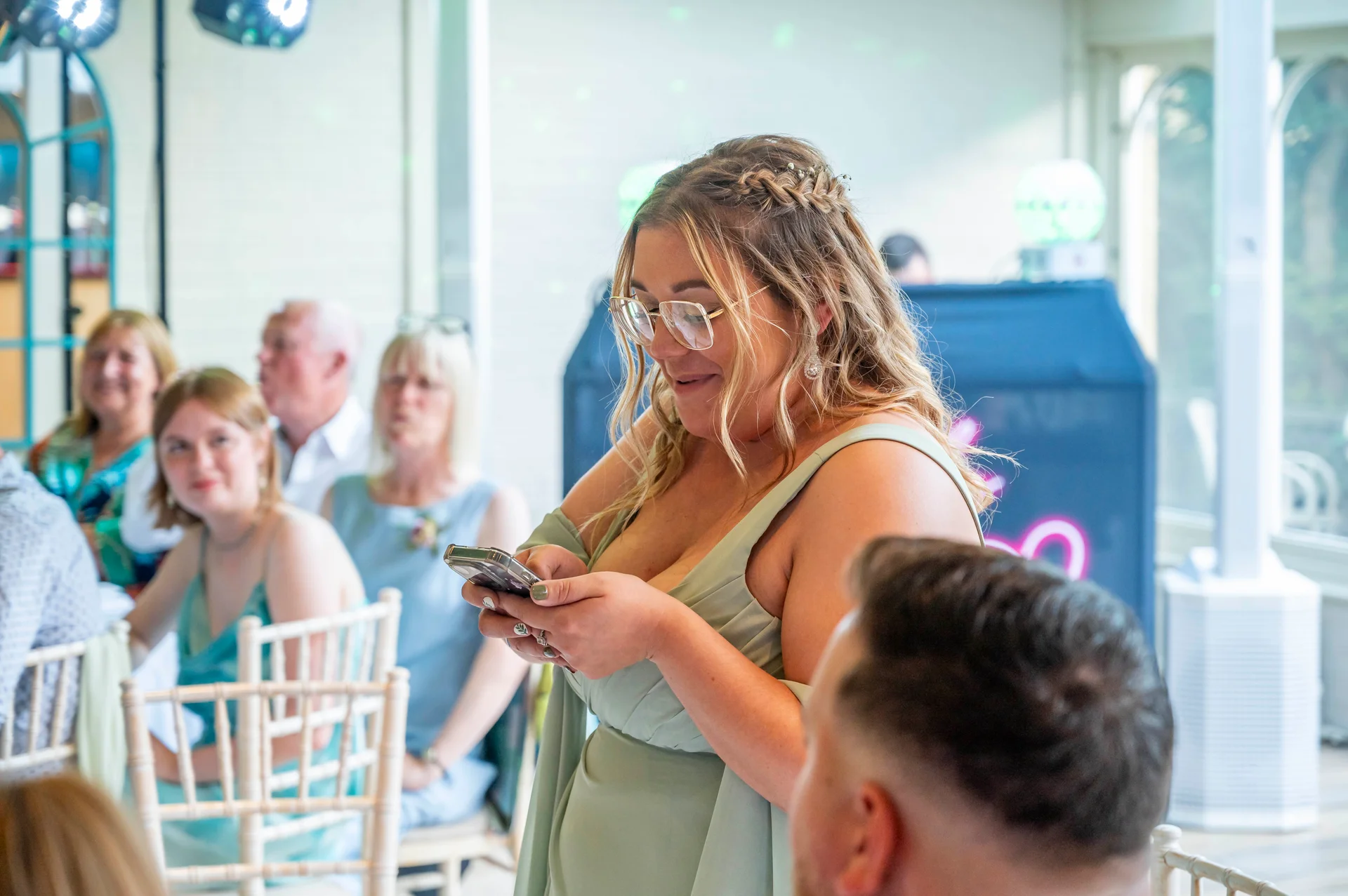 A guest wearing a sage green dress and glasses smiling down at her mobile phone during the wedding reception.