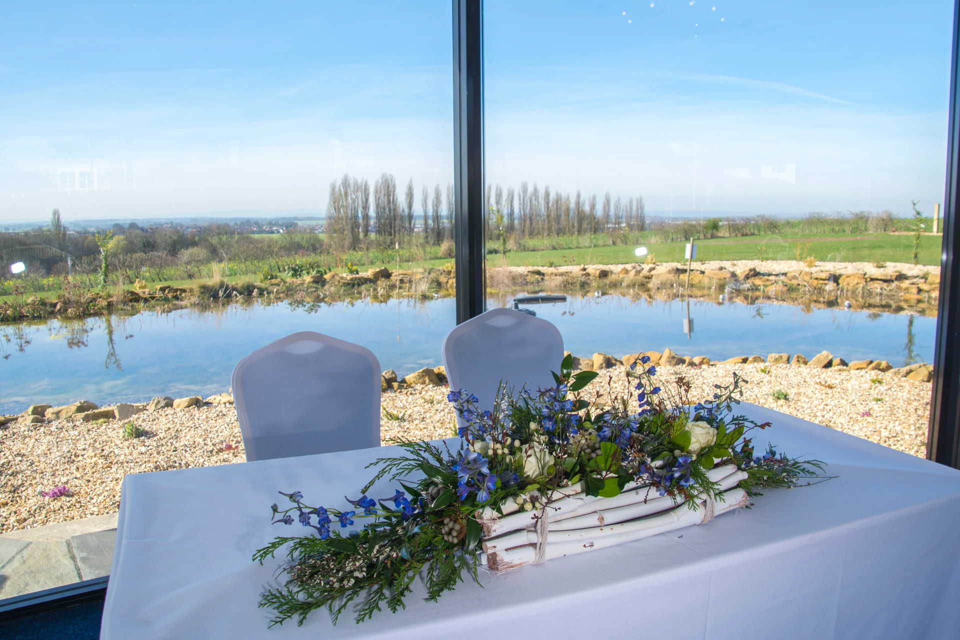 A elegantly styled top table featuring a vibrant floral arrangement of white roses and blue delphiniums sits in front of expansive floor-to-ceiling windows overlooking a serene lake and countryside view.