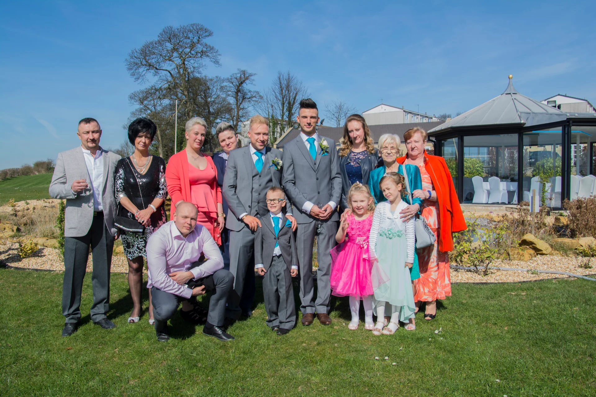 A large wedding group poses together outdoors on a manicured lawn under a clear blue sky on a sunny day.