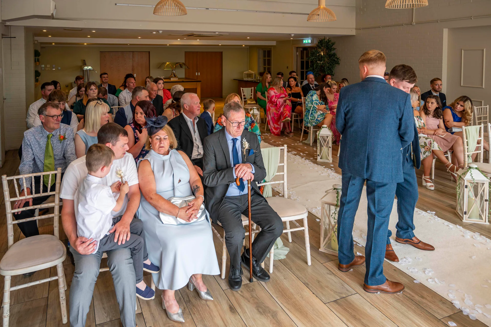 The groom and best man wait at the front of the ceremony room as seated guests watch from white chairs.