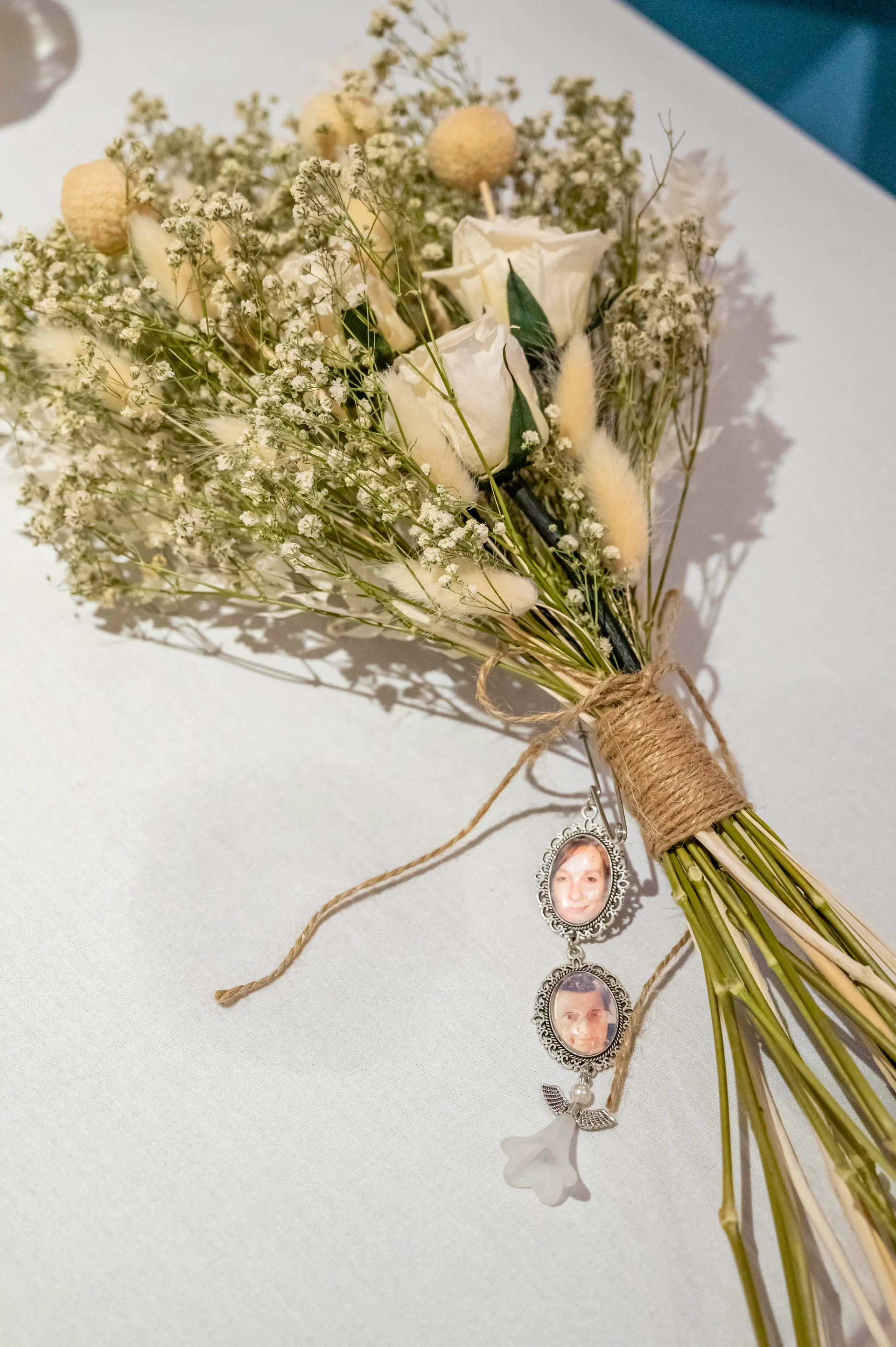 A close-up view of a hand-tied dried flower and white rose bouquet resting on a table, featuring two sentimental memorial photo charms tied to the twine-wrapped stem.