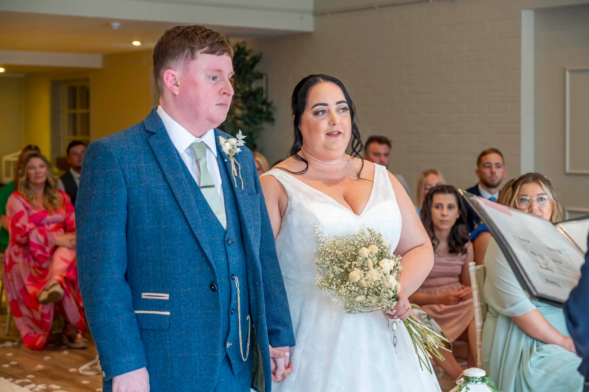 A bride and groom stand side by side during their civil ceremony, with the bride holding a textured bouquet of dried florals and the groom wearing a blue three-piece suit.