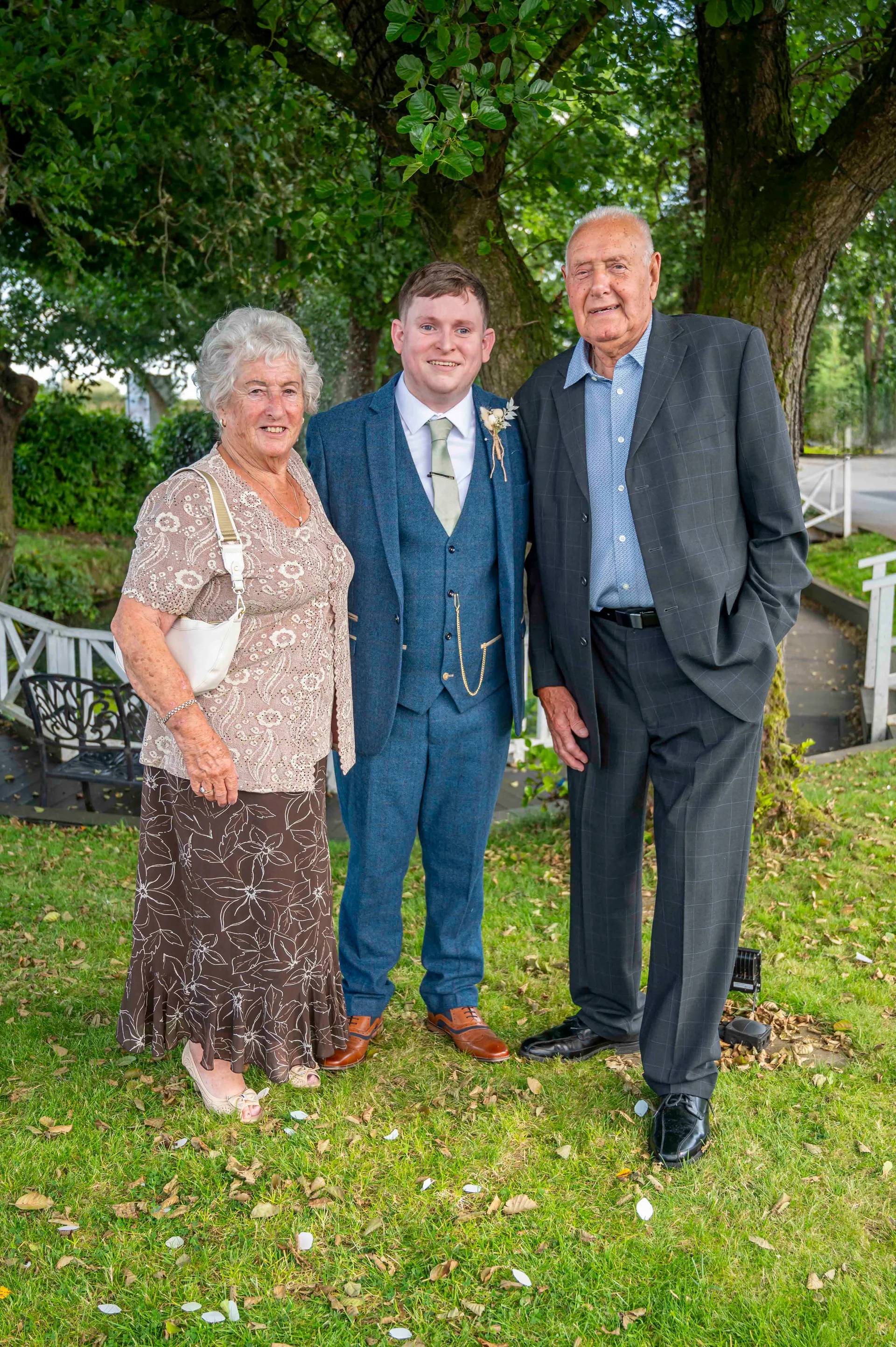 A groom in a blue tweed suit stands proudly flanked by his elderly grandparents outdoors on a grassy lawn beneath a mature tree.
