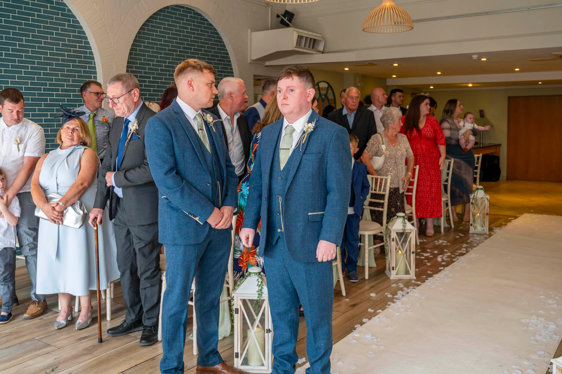 Two groomsmen in matching blue tweed three-piece suits stand at the front of a light-filled indoor ceremony space as they await the start of the wedding.