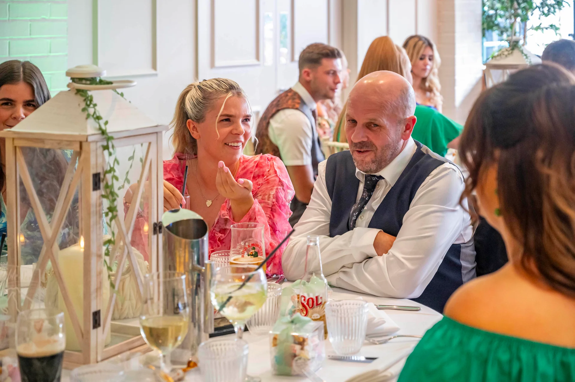 Guests laugh and converse cheerfully around a white-draped wedding breakfast table adorned with large decorative lanterns and candles.