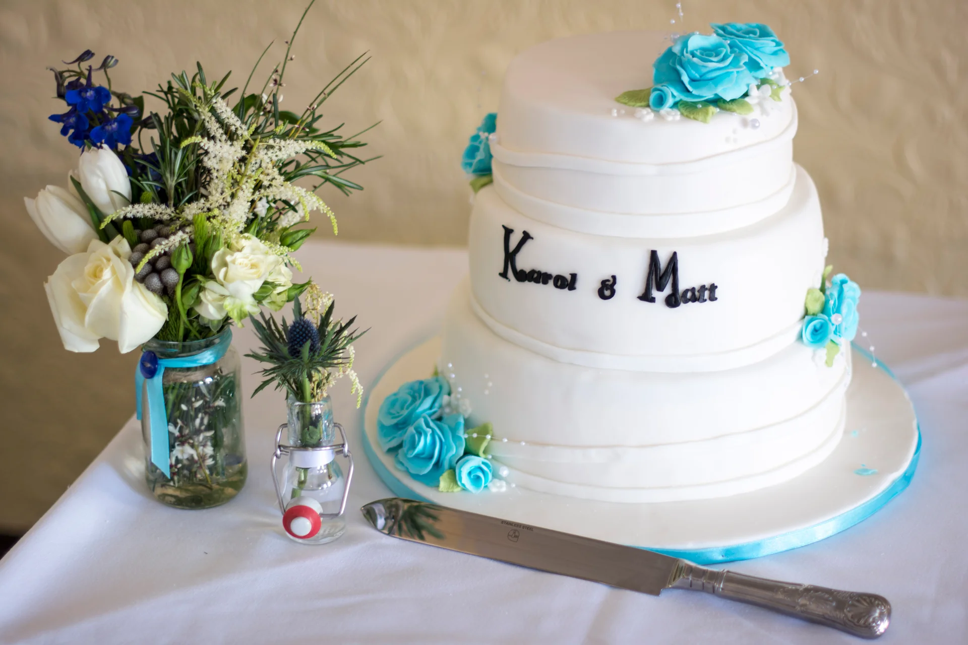 A three-tier white fondant wedding cake decorated with blue sugar roses sits next to a matching floral bouquet in a glass jar and a silver cake knife on a table.