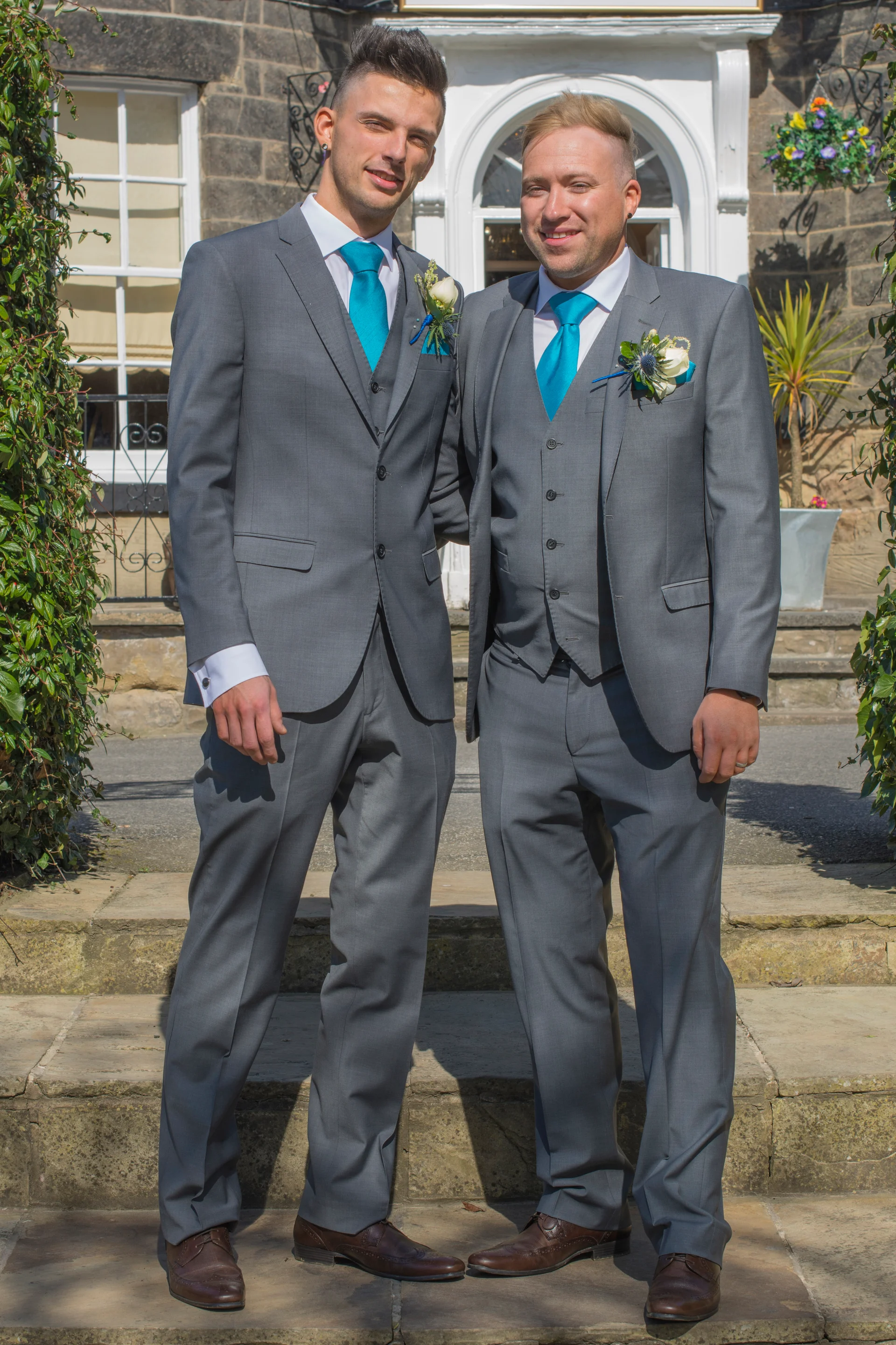 Two grooms in matching grey suits and bright turquoise ties pose together smiling outside a stone building.