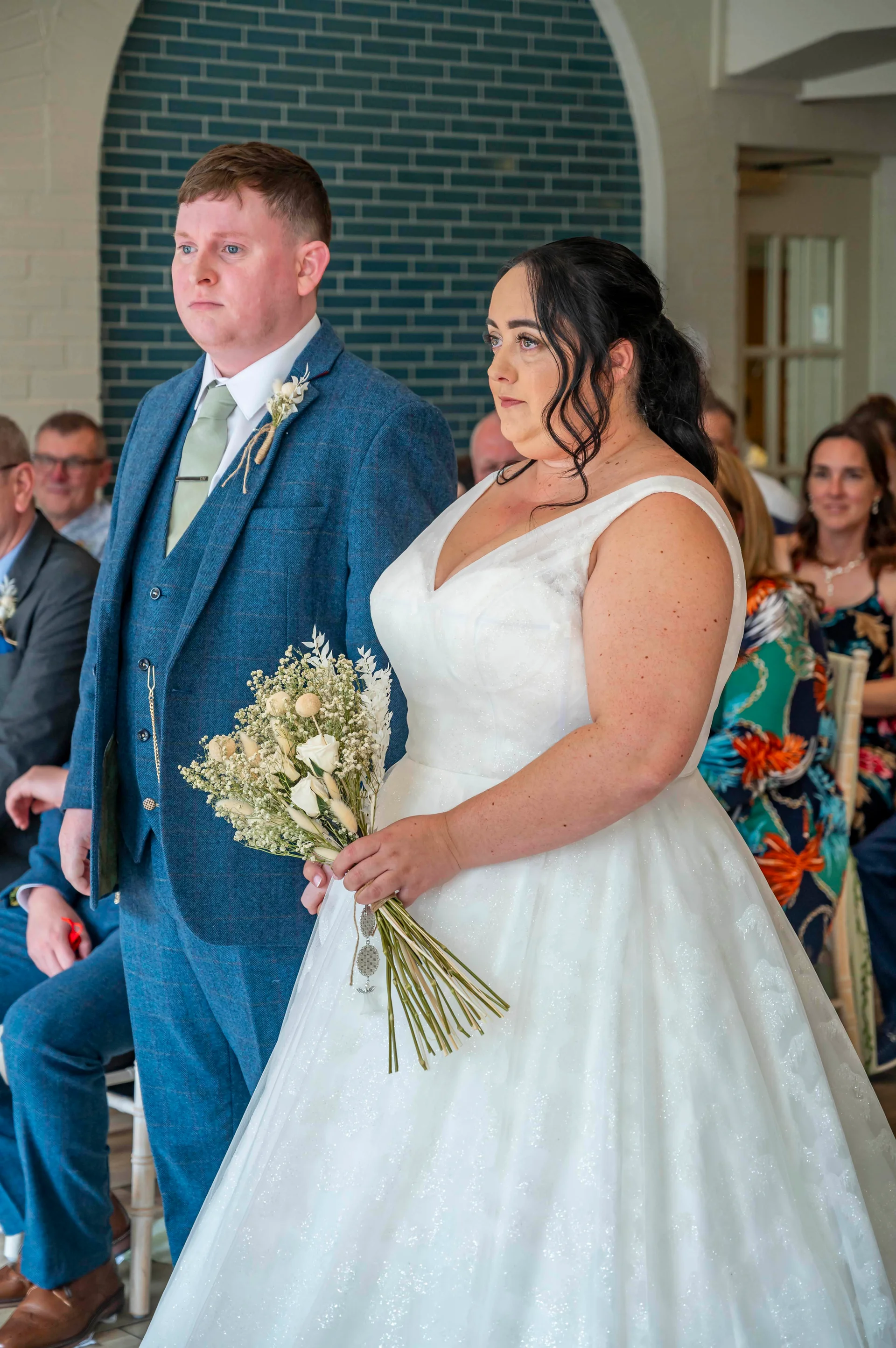 A newly married couple stands together during their ceremony, with the bride holding a bouquet of dried flowers and wearing a glittering A-line gown while the groom wears a blue tweed suit.