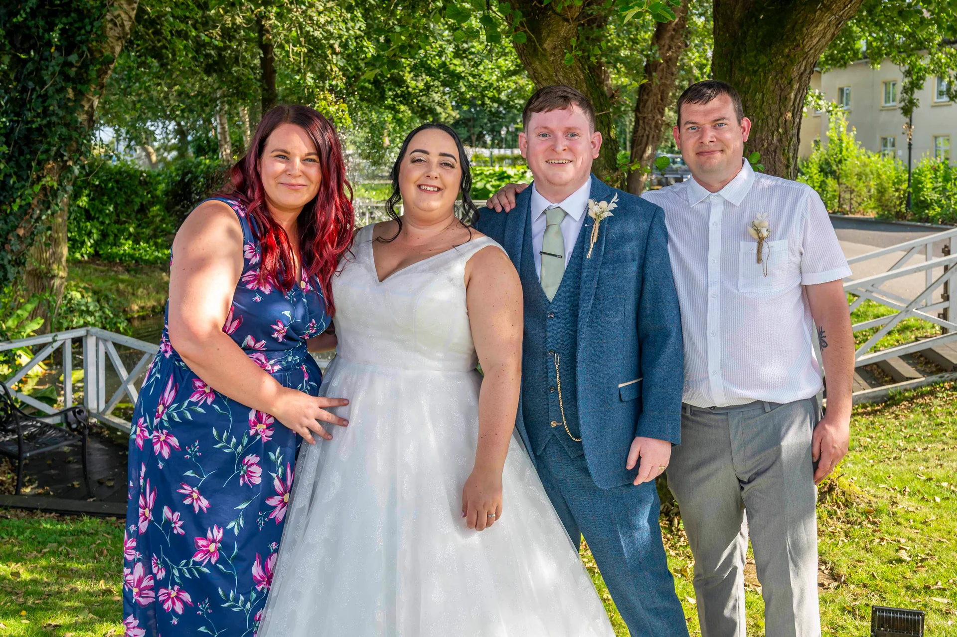 A bride in a white a-line gown stands alongside her groom in a blue tweed suit and two guests outdoors near a garden bridge.