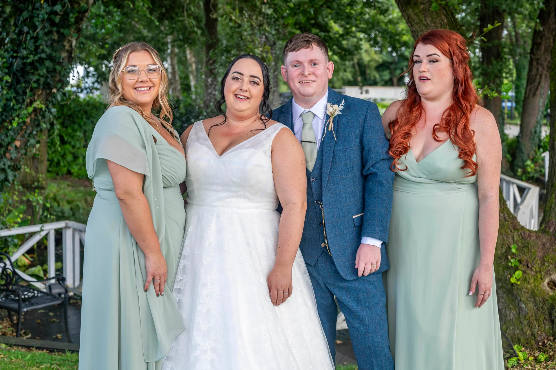 A smiling bride in a white a-line gown stands alongside a groom in a blue tweed suit and two bridesmaids wearing sage green dresses outdoors.