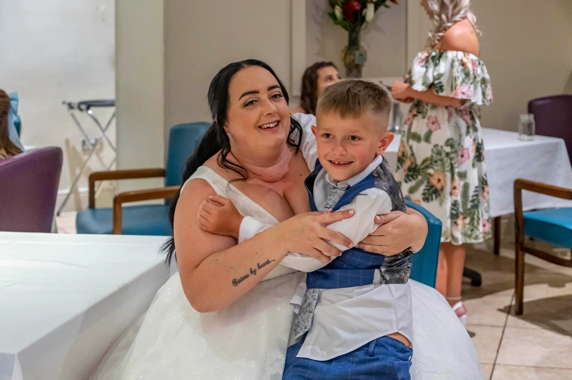 A joyful bride in a sparkly V-neck wedding dress warmly hugs a young page boy wearing a blue waistcoat indoors.
