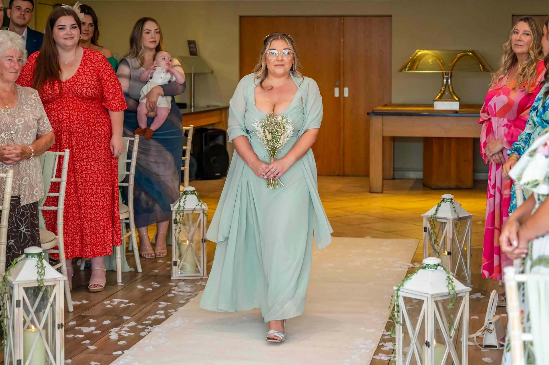 A bride in a sage green chiffon gown walks down a petal-strewn aisle carrying a bouquet of baby's breath, flanked by seated wedding guests and decorative white lanterns.
