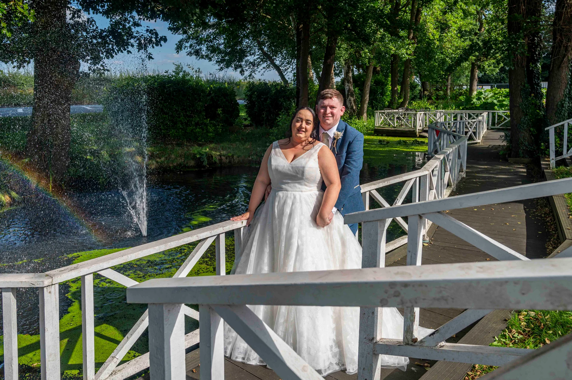 Newlyweds stand smiling together on a white wooden footbridge beside a pond featuring a fountain and a visible rainbow in the spray.