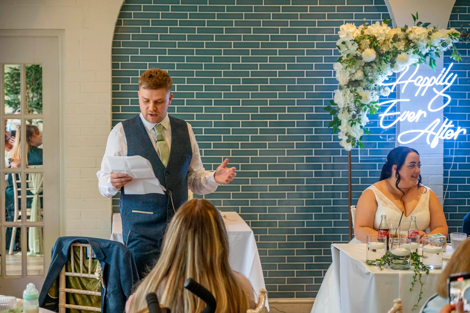 A groom delivers a speech holding notes in front of a blue tiled wall and illuminated neon sign, while a bride looks on from a reception table.