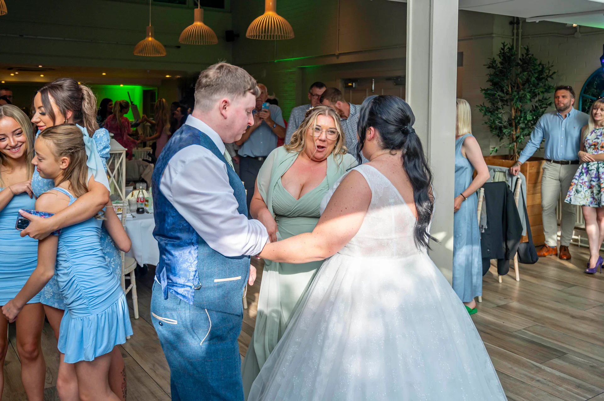 A newlywed couple shares a joyful dance during their reception, surrounded by smiling family and guests in a bright indoor venue with wooden flooring and pendant lights.