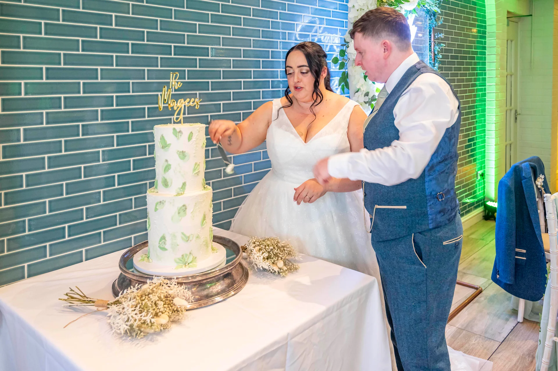 A bride and groom cut a two-tier white cake decorated with painted floral patterns against a backdrop of teal tiled brick.