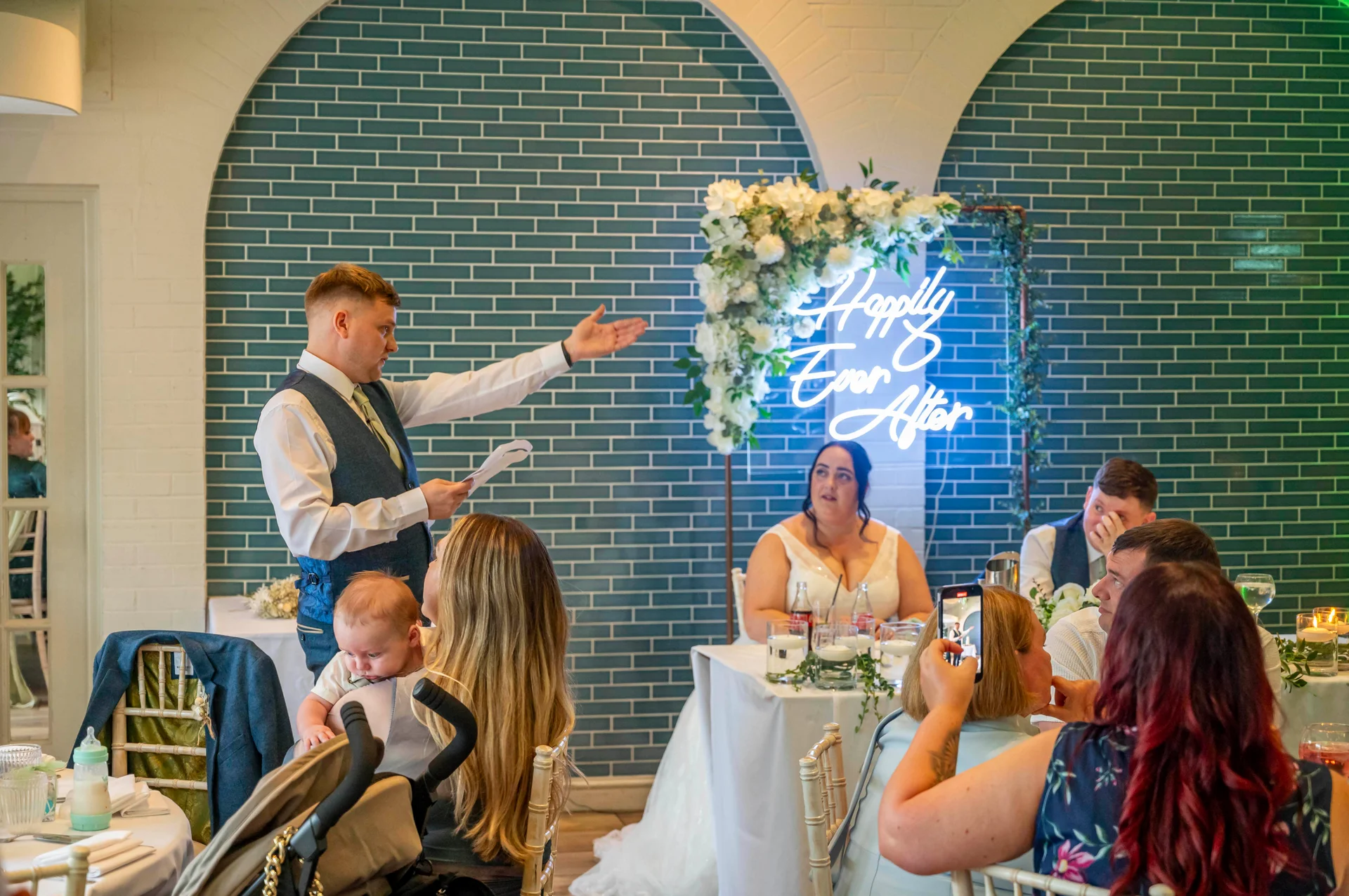A groomsman delivers an animated speech gesturing toward the head table while guests watch during the wedding reception.