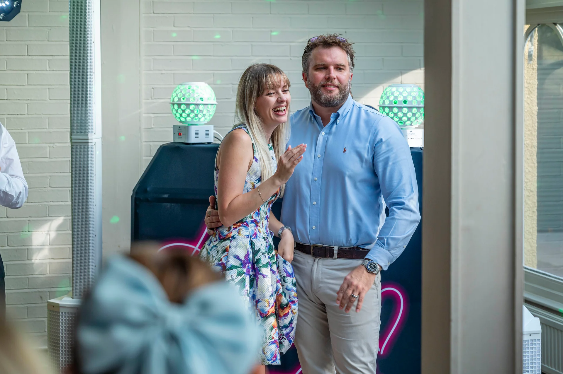 A smiling couple laughs joyfully together indoors against a white brick wall during a wedding reception.