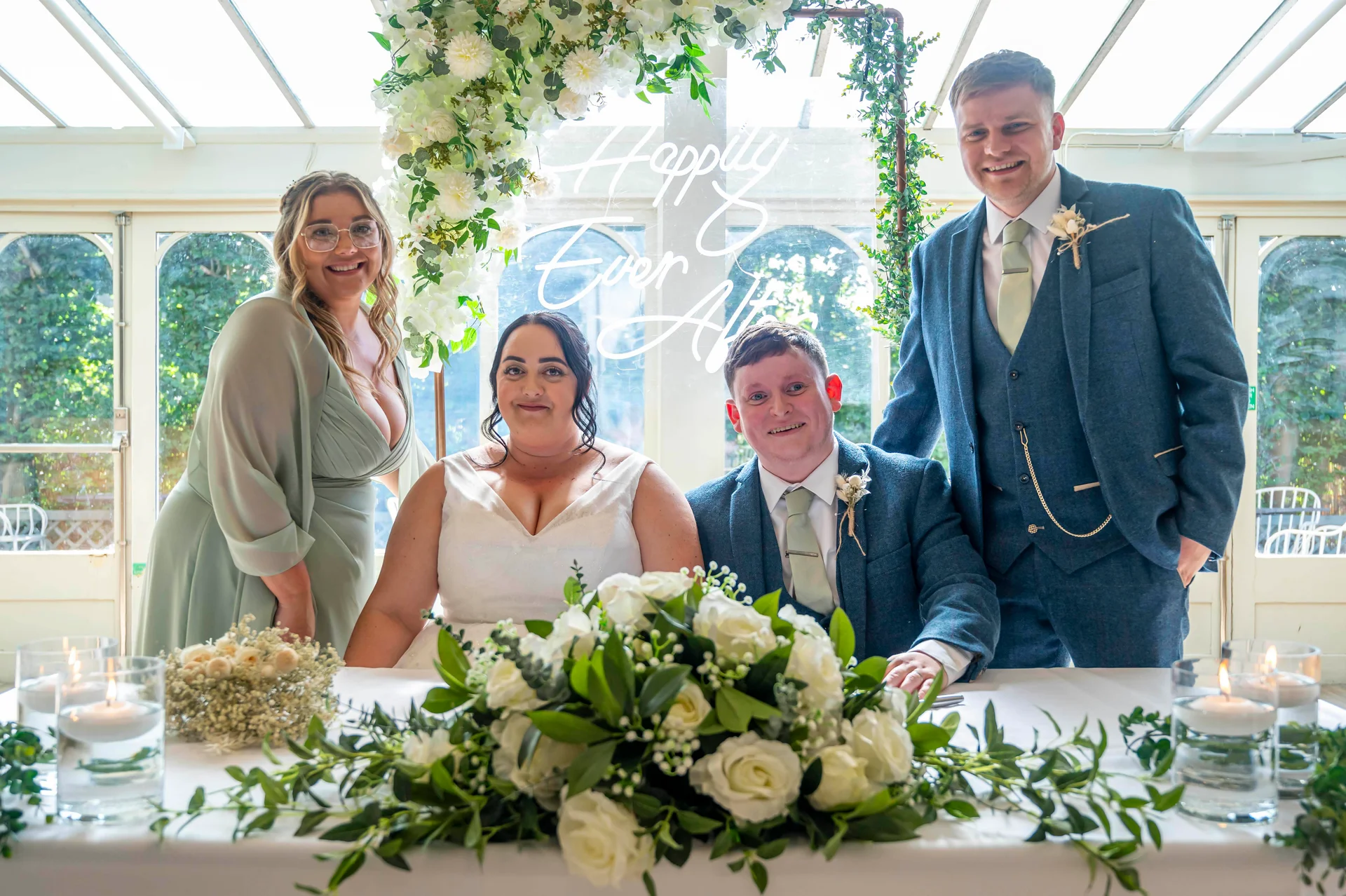 A newly married couple seated at a head table with a bridesmaid and groomsman in an orangery decorated with white florals and a neon sign.