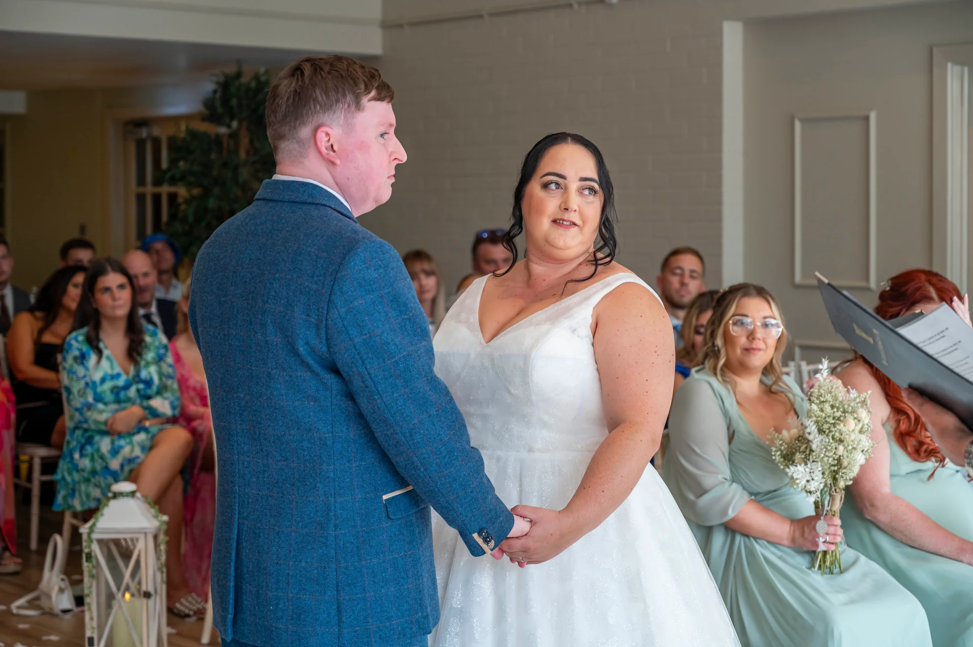 A bride and groom hold hands during their civil ceremony, with the groom wearing a blue tweed suit and the bride in a sparkling A-line wedding dress.